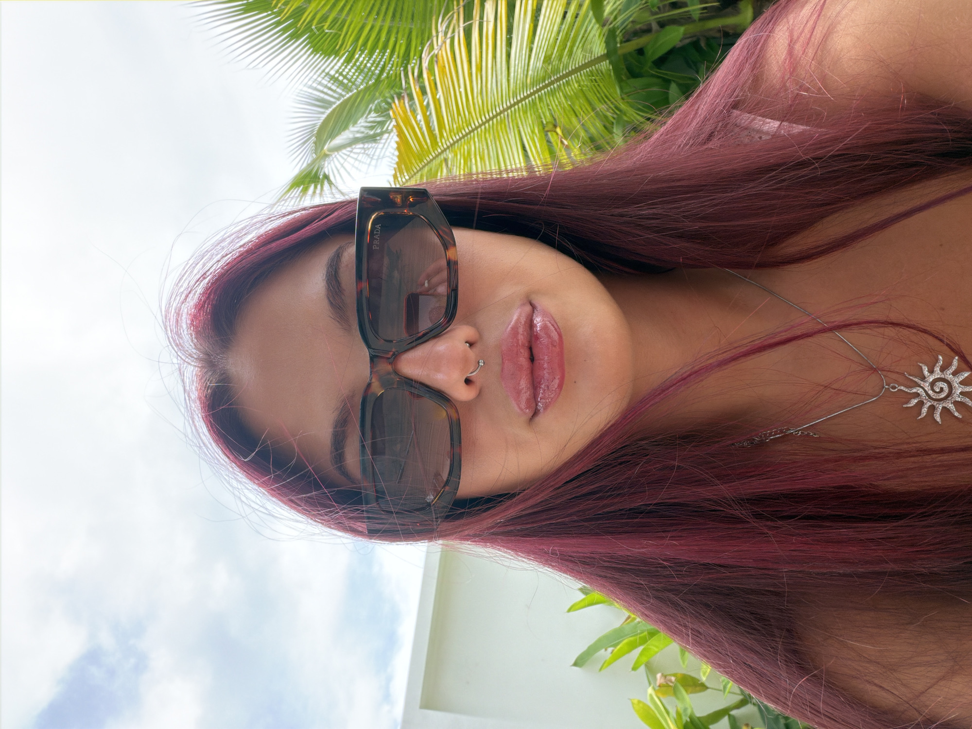 A close-up, selfie-style portrait of a young woman with long, dark pink hair and a nose ring. She is wearing large, tortoise-shell sunglasses with the "PRADA" logo visible on the side. Her lips are slightly parted and appear to have a glossy finish. She is wearing a silver necklace with a sun-shaped pendant. The background shows a bright sky with clouds, lush green palm fronds, and a white structure, suggesting an outdoor tropical setting. The overall lighting is bright, indicating daytime. The location is confirmed to be in the city of Choeng Thale, Thailand, given the characteristic tropical vegetation and architecture often found in resort areas.