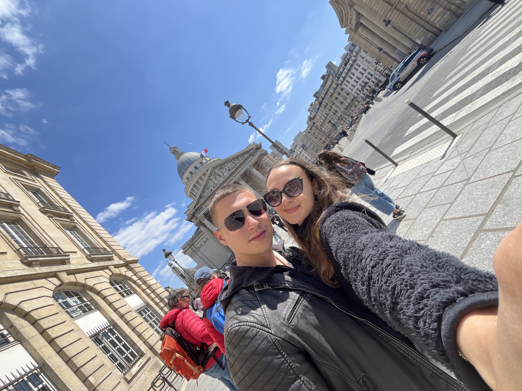 A wide-angle, slightly tilted selfie captures a young couple on a sunny day in Paris, France, with the iconic Panthéon building prominently in the background. The man, on the left, wears sunglasses and a black leather jacket over a dark hooded top. The woman, on the right, also wears sunglasses and a dark, fuzzy jacket, with her arm extended to take the photo. Both appear relaxed, and the woman has a slight smile.
The setting is an urban street scene under a clear, bright blue sky with scattered white clouds. The grand, domed structure of the Panthéon, featuring classical columns and a pediment, occupies the background, with a small French flag visible near its dome. To the left, a multi-story classical Parisian building with ornate windows and balconies is partially visible.
In the middle ground, a traditional Parisian street lamp stands tall. Pedestrians can be seen walking around, including an individual in a red jacket and an orange backpack. Further back, a street features a clear zebra crossing and a parked French police car, distinguishable by its white, blue, and red markings and the word "POLICE" (partially visible in crops). The ground is paved with square tiles. Partially visible text "MAIRIE DE" can be discerned on a building in the background. The strong sunlight casts clear shadows, indicating midday or early afternoon.
