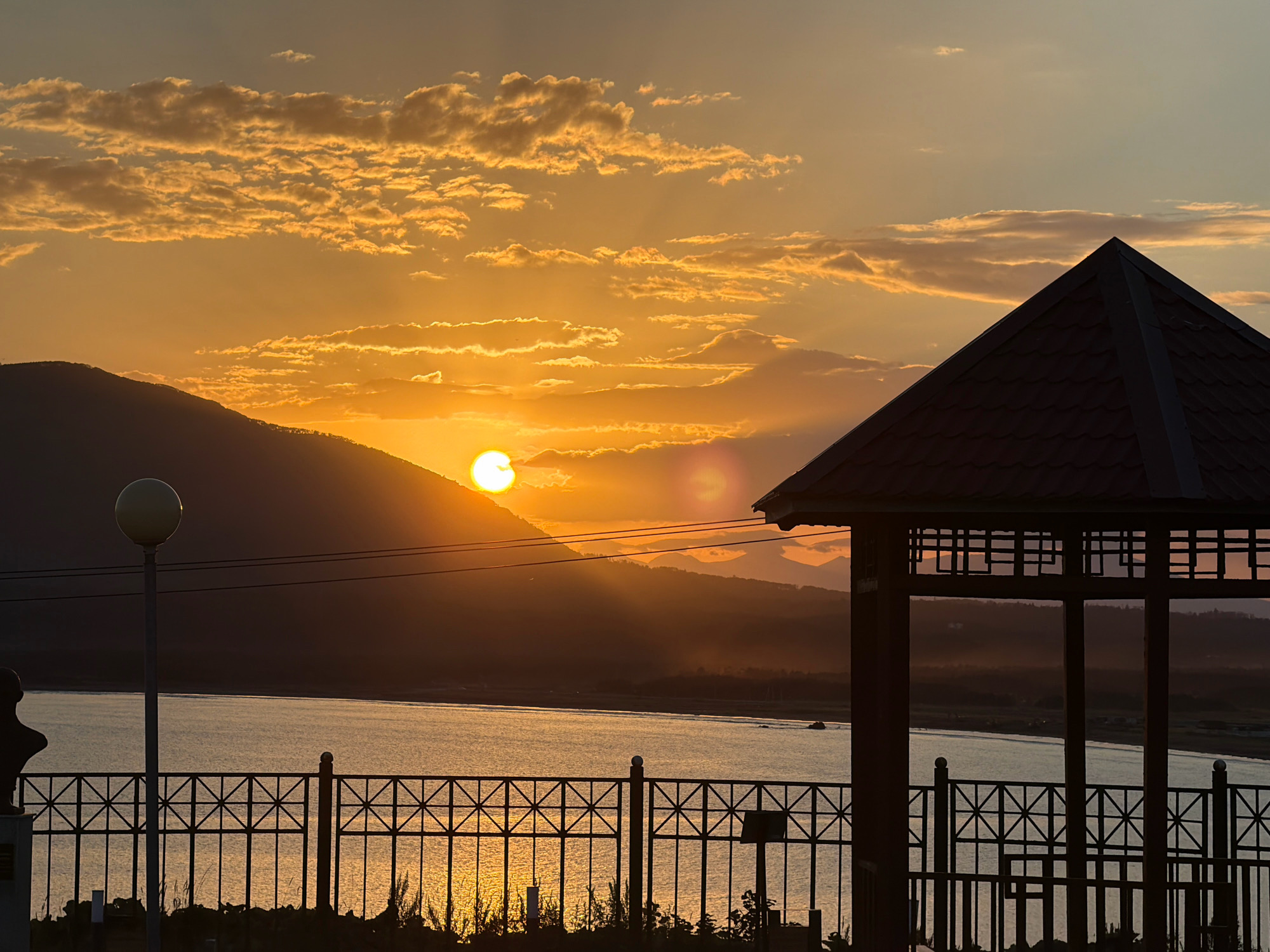 A sunset casts a warm golden glow over a tranquil body of water, reflecting the sky's fiery hues. Silhouetted against the vibrant sky are rolling hills and mountains, their dark forms creating a dramatic contrast. In the foreground, a dark, ornate gazebo with intricate latticework stands on the right, its roof a sharp pyramid against the horizon. To the left, a black metal fence with a geometric pattern separates the viewer from the water. A lamppost with a spherical light fixture rises beside a dark bust on a pedestal, also silhouetted. The scene is set in Yuzhno-Kurilsk, Russia, a coastal town known for its stunning natural beauty. The water is calm, with a distant boat visible, leaving a faint wake on its surface. The overall atmosphere is one of serene natural beauty, captured at the end of a day.