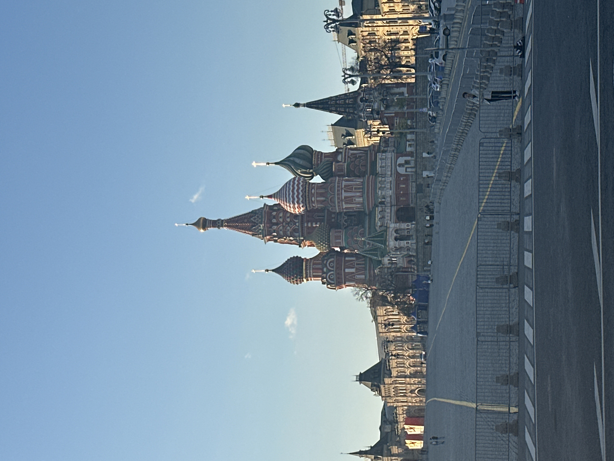 The image shows St. Basil's Cathedral in Moscow, Russia. The cathedral's distinct, multi-colored onion domes are prominently visible against a clear blue sky. Adjacent to the cathedral is Red Square, part of which is visible. A street runs along the edge of the square, marked with road lines and barriers. Buildings are seen in the background. A person is seen walking along the road.
