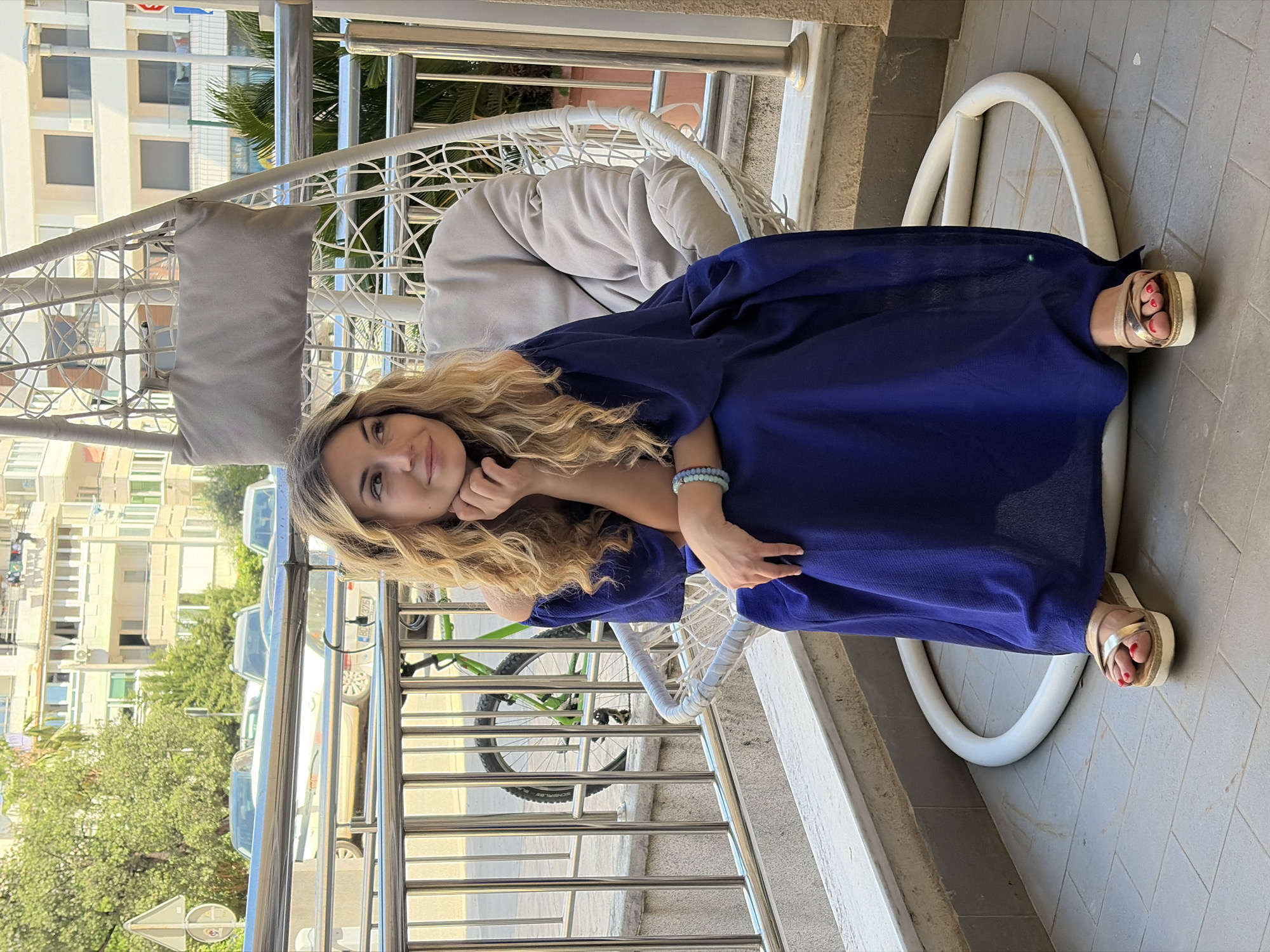 A woman is seated in a white egg-shaped chair on a balcony. She is wearing a long, flowing blue dress and gold sandals. Her blonde hair is wavy and falls around her shoulders. She is holding her hand to her face and looking towards the camera. The balcony has a silver metal railing and a white edge. A green bicycle is visible behind the railing. In the background, there are apartment buildings, trees, and parked cars, characteristic of the city of Budva, Montenegro. The image suggests a relaxed, outdoor setting in a residential area.