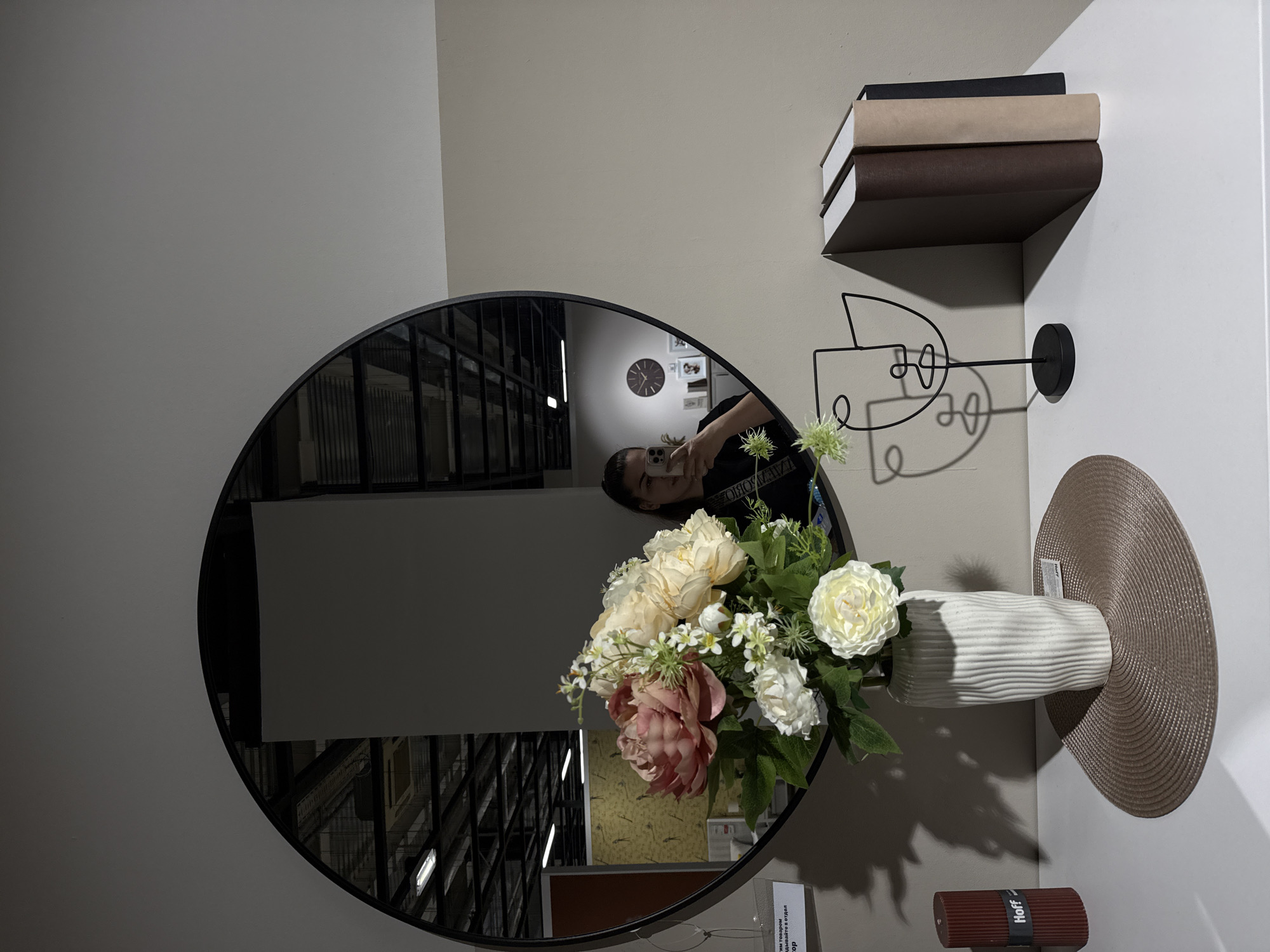 A round mirror hangs on a light beige wall, reflecting the interior of a store. In the foreground, a white vase with a textured surface holds a bouquet of artificial flowers, predominantly pink and cream peonies, with smaller white blossoms and green foliage. The vase sits on a round, woven placemat on a white surface. To the left of the vase is a dark red candle with fluted sides, bearing the "Hoff" logo. To the right of the vase, a minimalist black metal sculpture of a face stands. Behind the sculpture, three books with dark brown, tan, and black covers are propped upright.
The mirror's reflection shows a woman holding a phone, taking a selfie. She is wearing a black t-shirt with white lettering. In the background of the reflection, the store's ceiling structure with lights and shelves is visible. A large clock with Roman numerals is also visible in the reflected background. There is no specific information in the image that allows for determining the location in Krasnoyarsk, Russia.