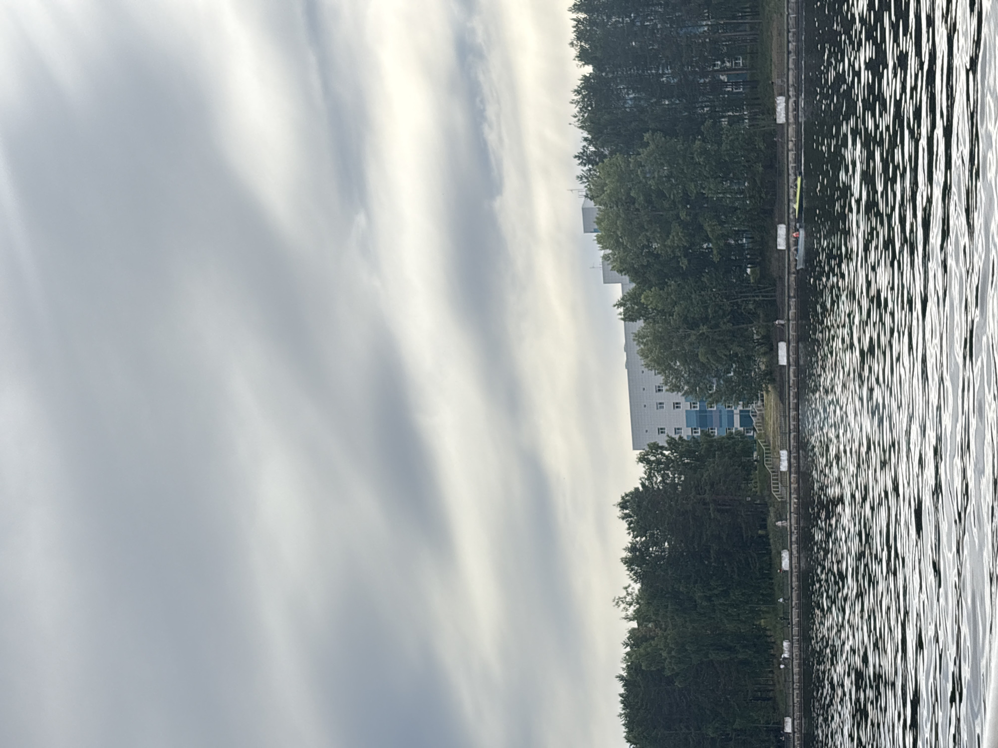 The image depicts a wide shot of a body of water, likely the Kama River or one of its tributaries, in Chaikovsky, Russia. The water's surface is rippled, reflecting the overcast sky and creating a textured pattern.
In the background, a tree-lined embankment runs parallel to the water. A paved walkway is visible atop the embankment, with several individuals walking or standing along it. Beyond the trees, a multi-story building with a white facade and blue-tinted windows is partially visible. The building appears to be an institutional or residential structure.
On the water, a small boat is moving, carrying at least two individuals. The boat is light-colored, and one of the occupants is wearing an orange life vest. The scene suggests a calm, possibly late afternoon or early morning setting given the diffused lighting and cloud cover.