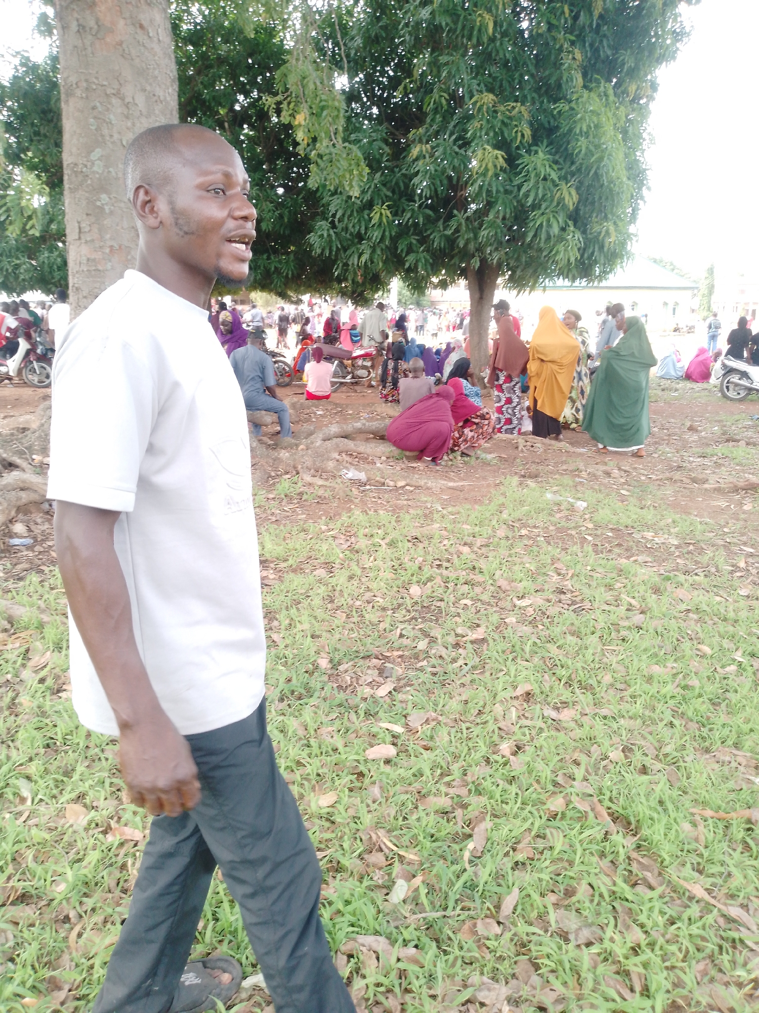A man in a white t-shirt and dark pants walks toward the right in a grassy area with a tree to his left. In the background, a large group of people, many wearing traditional robes, are gathered in a dusty area with motorcycles nearby. The location appears to be an open space in the city of Unknown, Unknown.