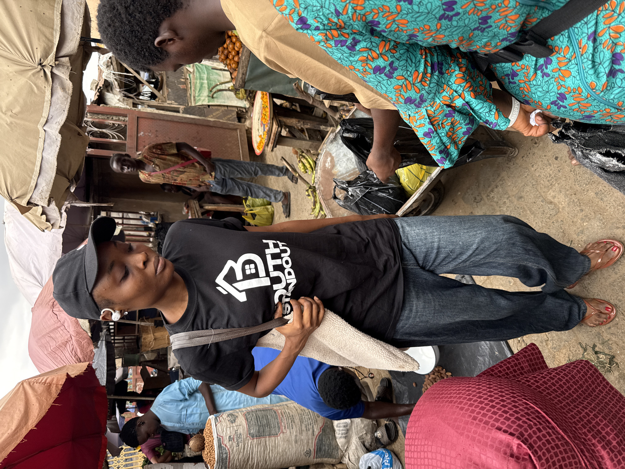 The image depicts a crowded marketplace in Lugbe, Nigeria. A woman wearing a black t-shirt, blue jeans, a baseball cap, and wireless earbuds stands out in the foreground. She carries a grey shoulder bag and appears to be interacting with vendors or shoppers. The market is bustling with people, many of whom are buying or selling goods. Merchandise includes bananas, other fruit, and what appears to be sacks of grains. The stalls are covered with colorful awnings. The surroundings show the typical infrastructure of a busy African marketplace.