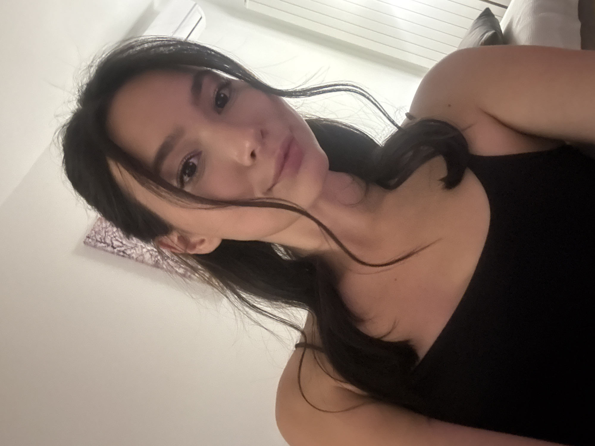 A close-up, angled shot shows a woman with long, dark hair, partially pulled back, wearing a black tank top. The background includes a white wall, what appears to be an air conditioning unit, and a glimpse of a pillow in neutral tones. The woman's face is the focal point, with a slight smile.