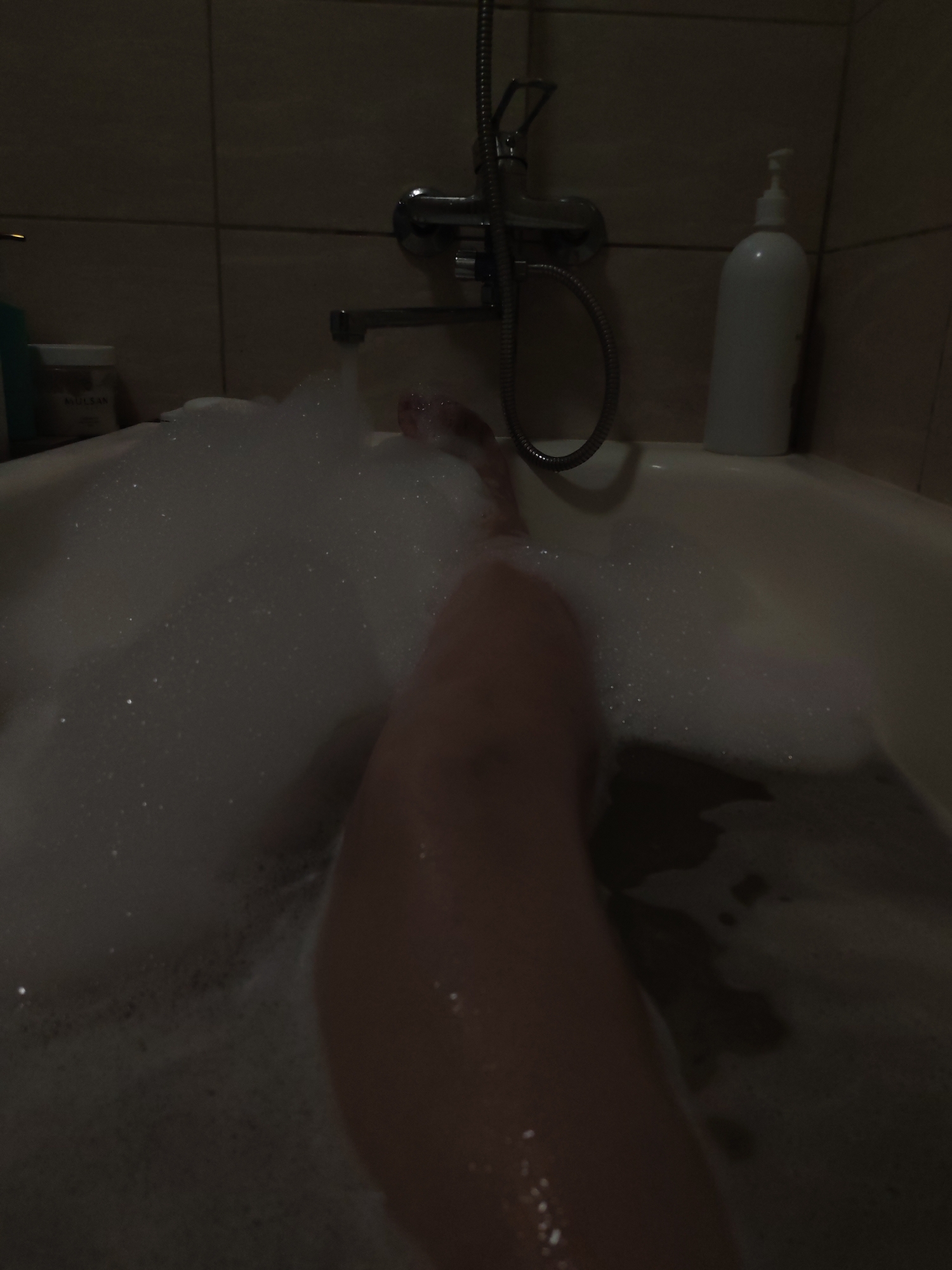 The image shows a partially-filled bathtub with soapy water and bubbles. A person's leg is visible in the tub. In the background, there is a tile wall, a faucet, and a bottle of lotion. The location is a bathroom. There is no information to indicate this is in Izhevsk, Russia.