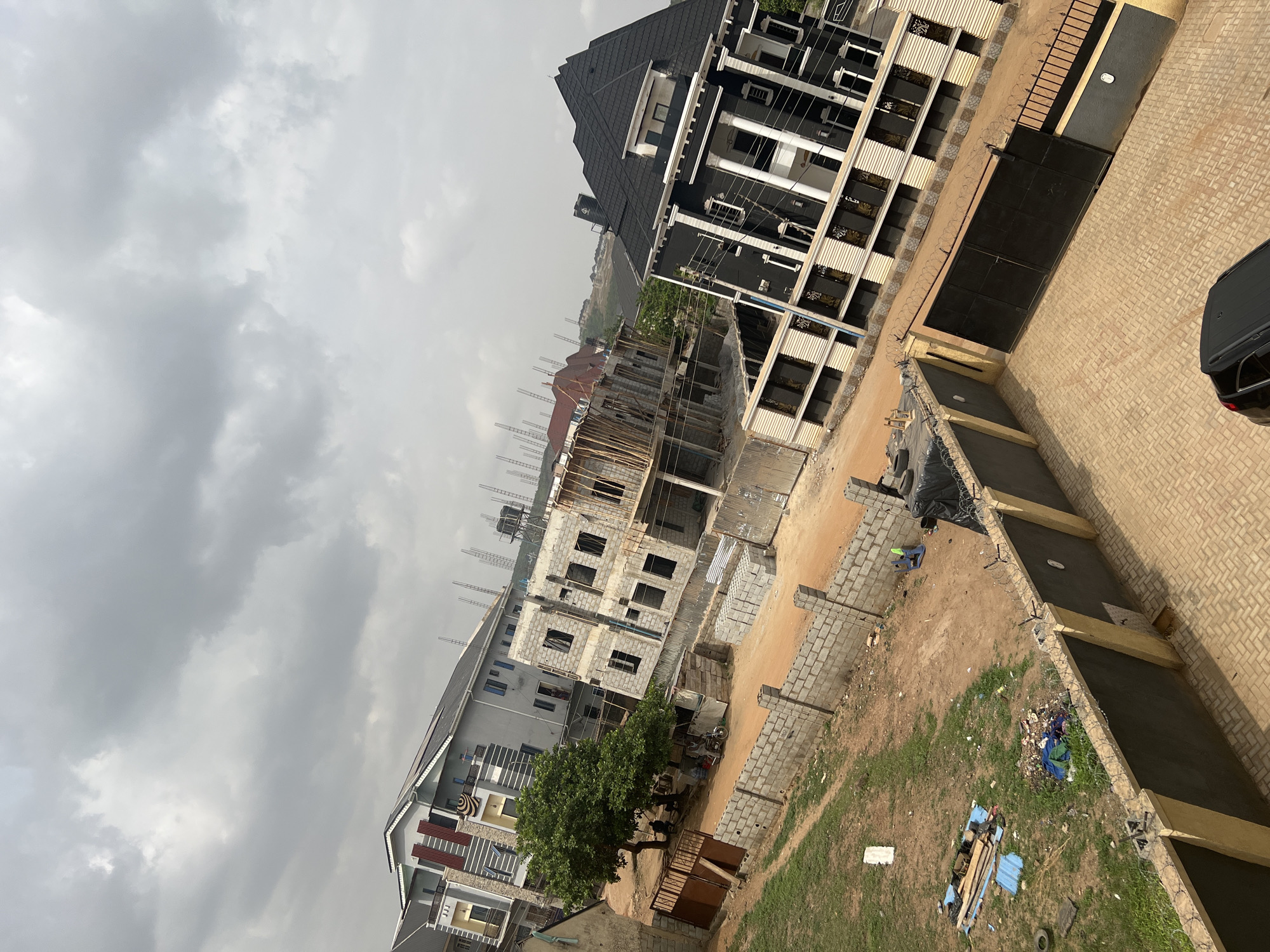 The image shows an urban landscape in Abuja, Nigeria, featuring multiple buildings in varying states of completion and occupation. The sky is overcast with heavy, grey clouds.
In the foreground and midground, there are several structures under construction. These include exposed concrete and scaffolding, indicating ongoing building activity. One partially-constructed building has a wooden framework surrounding it.
Completed buildings are also visible. One has a modern design with a black roof and white pillars. Another completed building has a grey exterior and multi-colored accents on the facade.
A patch of land is visible to the right. Litter and construction debris are scattered in this area. The ground appears a mixture of dirt and grass, separated by brick edging, with discarded boards, metal, and plastic.
The streets appear to be sandy or unpaved near construction areas. A vehicle, partially obscured on the right edge, is visible.
The presence of scaffolding, incomplete buildings, and construction materials suggests this is a developing or recently developed part of the city.