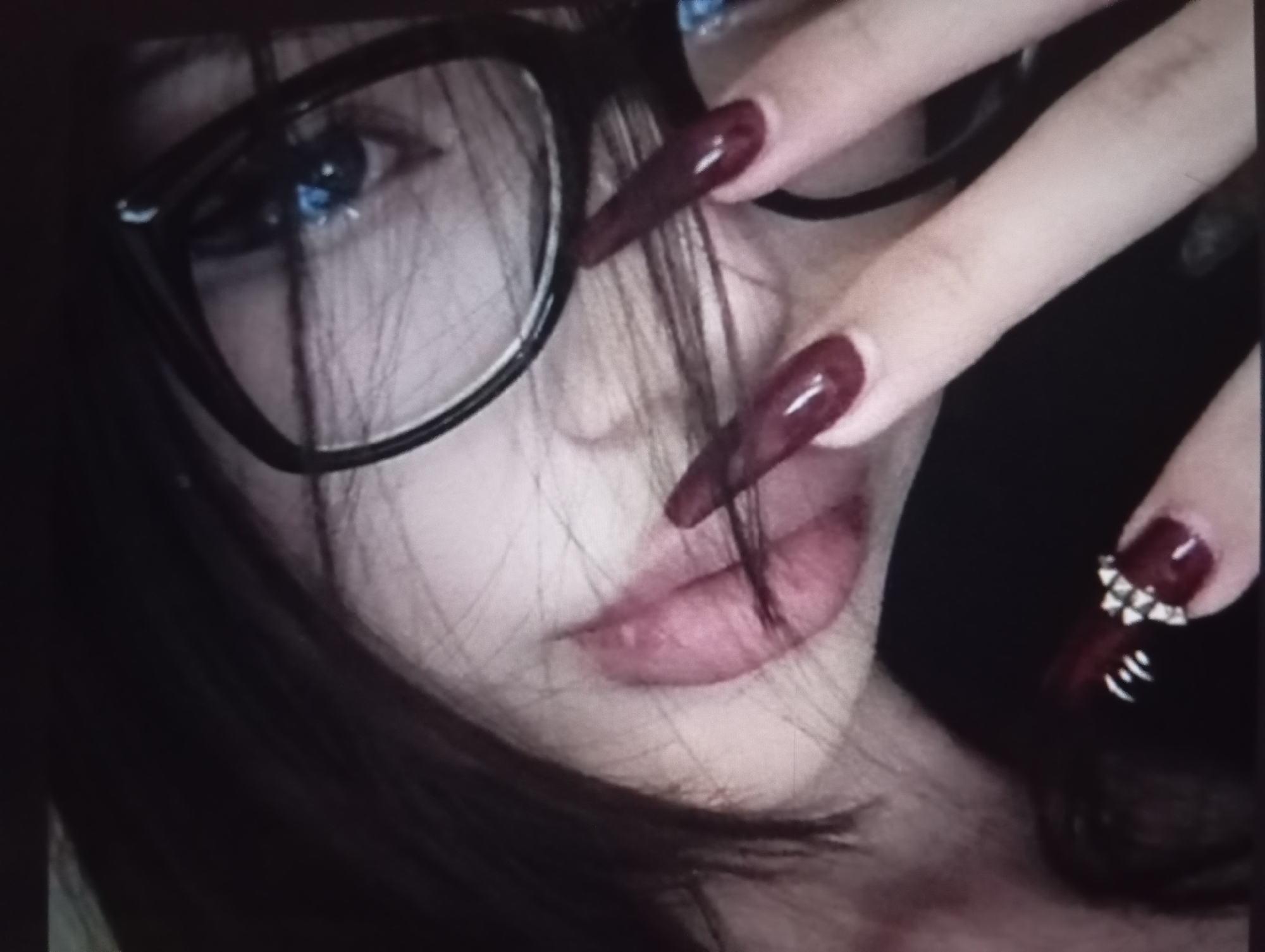 A close-up shot features a person with fair skin, dark hair partially obscuring the face, and wearing large, black-rimmed glasses that frame a bright blue eye. The focus is on the face and the hand strategically placed to further obscure the facial features. The fingernails are long, pointed, and painted in a dark red color, with one nail adorned with silver studs and lines. The lips are visible, and the overall impression is one of posed obscurity. There is no information to suggest this image was taken in Odesa, Ukraine. The image only contains a close-up shot of an individual and lacks any identifiable contextual information about the location.