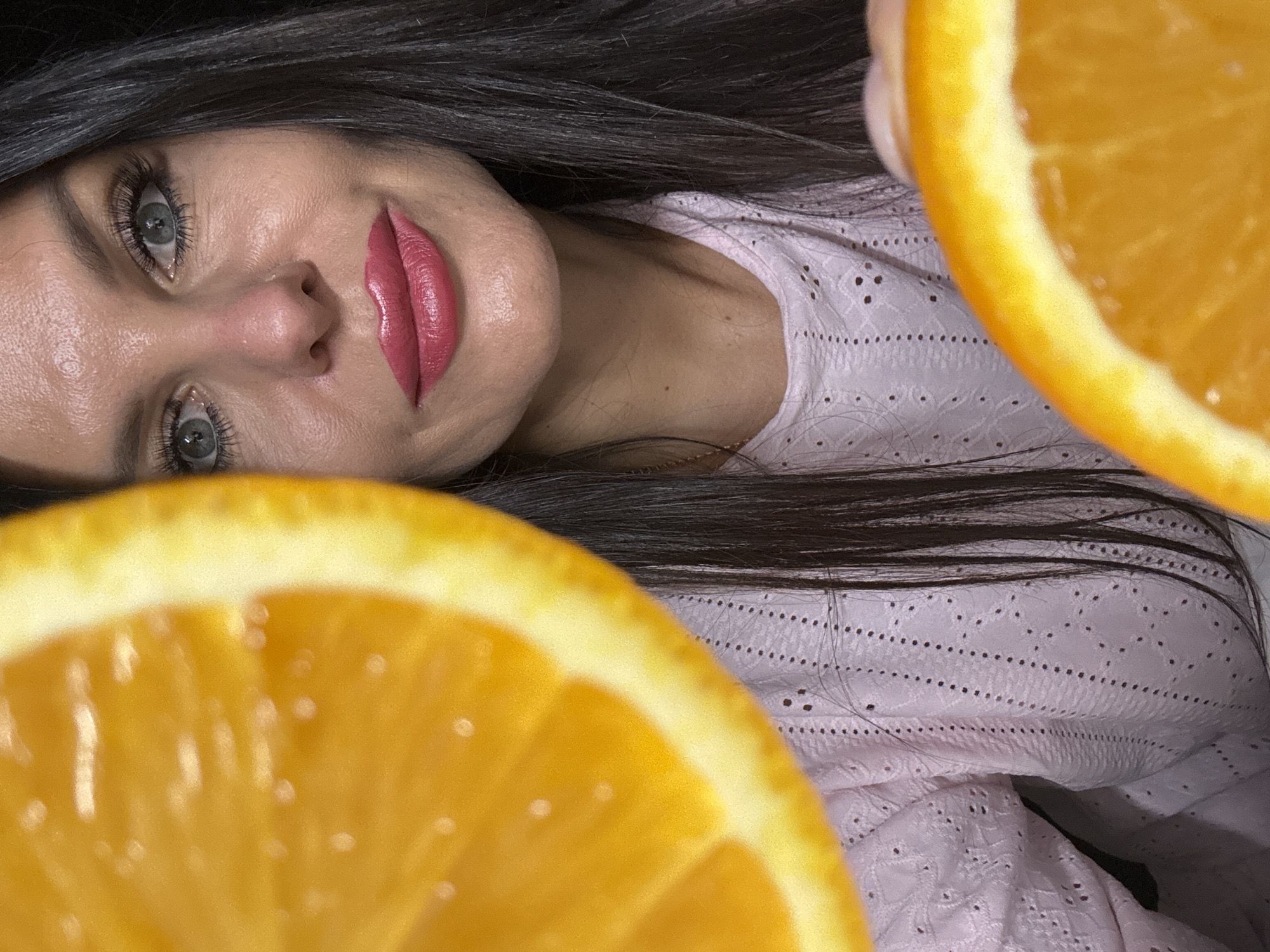 A woman with dark hair and light eyes lies on her side, facing the camera. She wears a light pink eyelet shirt. Two orange halves frame her face. The location, Chaikovsky, Russia, cannot be confirmed.