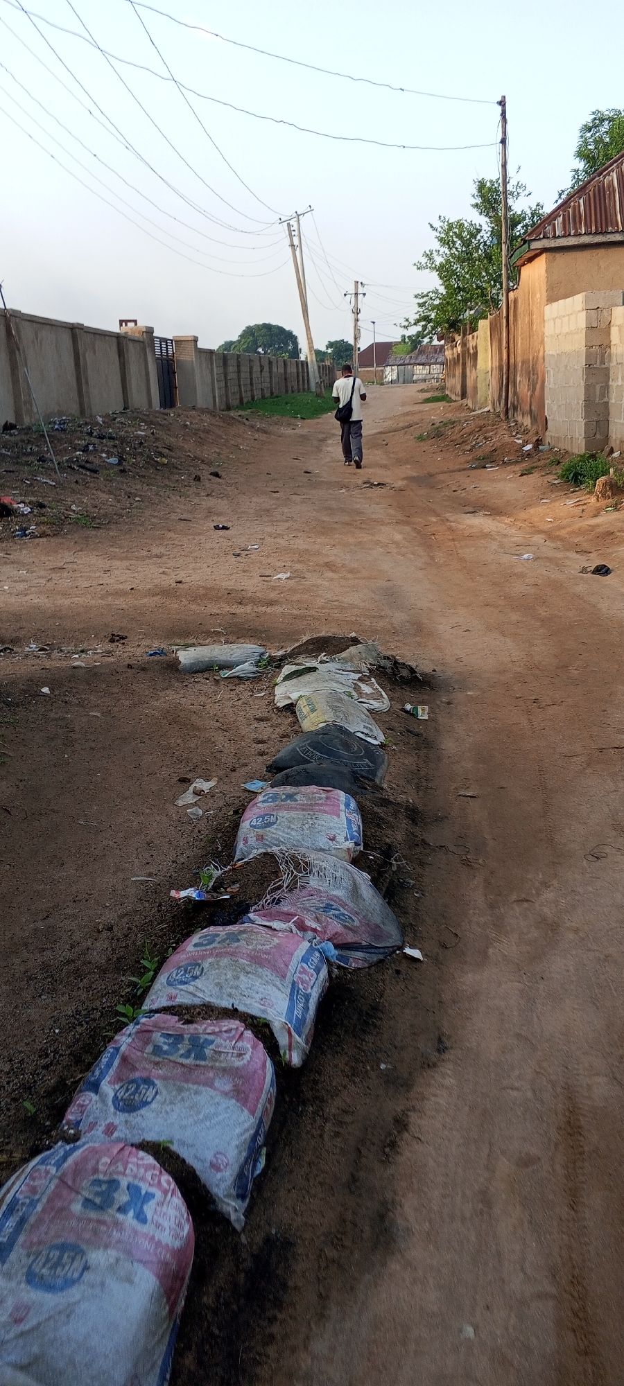A man walks down a dirt road in Jalingo, Nigeria, toward buildings and trees in the distance. He is wearing a light-colored shirt and dark pants, carrying a dark bag over his shoulder. The road is lined with sandbags filled with dirt, and there is scattered debris and trash along the sides. Concrete walls and fences border the road in some areas, and power lines crisscross overhead. The sky is overcast.