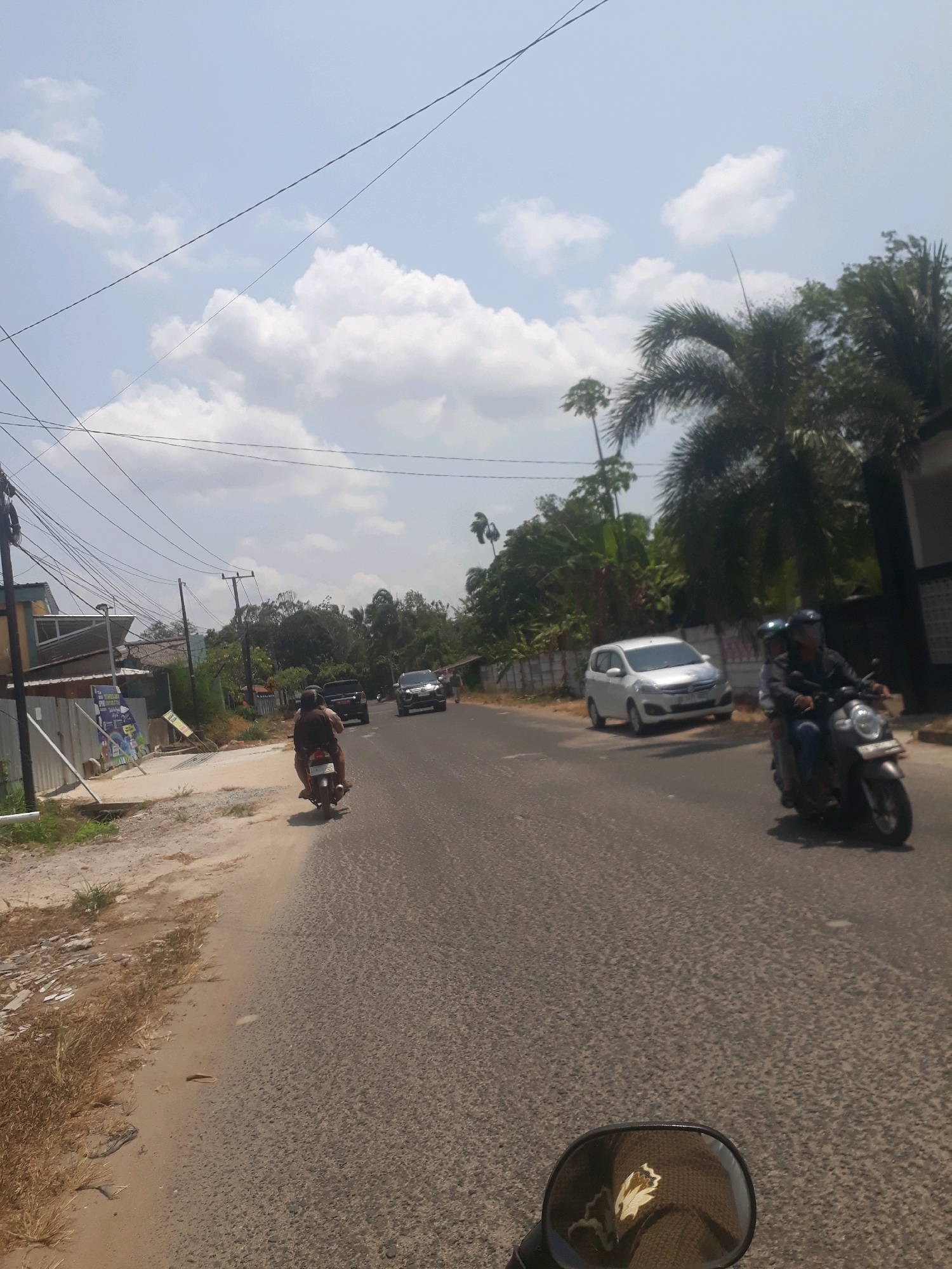 The image shows a street scene in the city of Pangkalpinang, Indonesia, on a sunny day with scattered clouds. In the foreground, a motorcycle rearview mirror reflects a brown patterned object. The road, made of asphalt, is wet in places and has visible texture. On the right side of the road, a motorcycle with two riders, both wearing helmets, is moving forward. The rider in front is wearing a dark jacket and blue pants, while the passenger is wearing a brown shirt. On the left side of the road, a person is riding a motorcycle, also wearing a helmet and a brown shirt. Further down the road, a white car is parked on the side, and two other cars, a black SUV and a silver car, are driving in the same direction as the motorcycles. The roadside is lined with vegetation, including palm trees and other green foliage. On the left, there are some buildings and a fence. Overhead, power lines crisscross the sky. The overall impression is a typical street scene in a tropical urban environment.