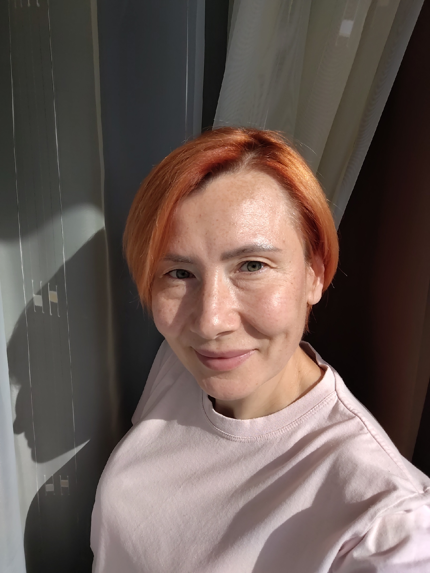 A woman with short, reddish-orange hair and fair skin with freckles is looking at the camera in a selfie. She is wearing a light pink t-shirt. The background consists of vertical blinds and sheer curtains. A shadow of the woman's head is cast on the blinds to her left. The lighting suggests it is daytime, with sunlight creating shadows and highlighting her face. The scene appears to be indoors, possibly in a home in Konakovo, Russia, given the context.