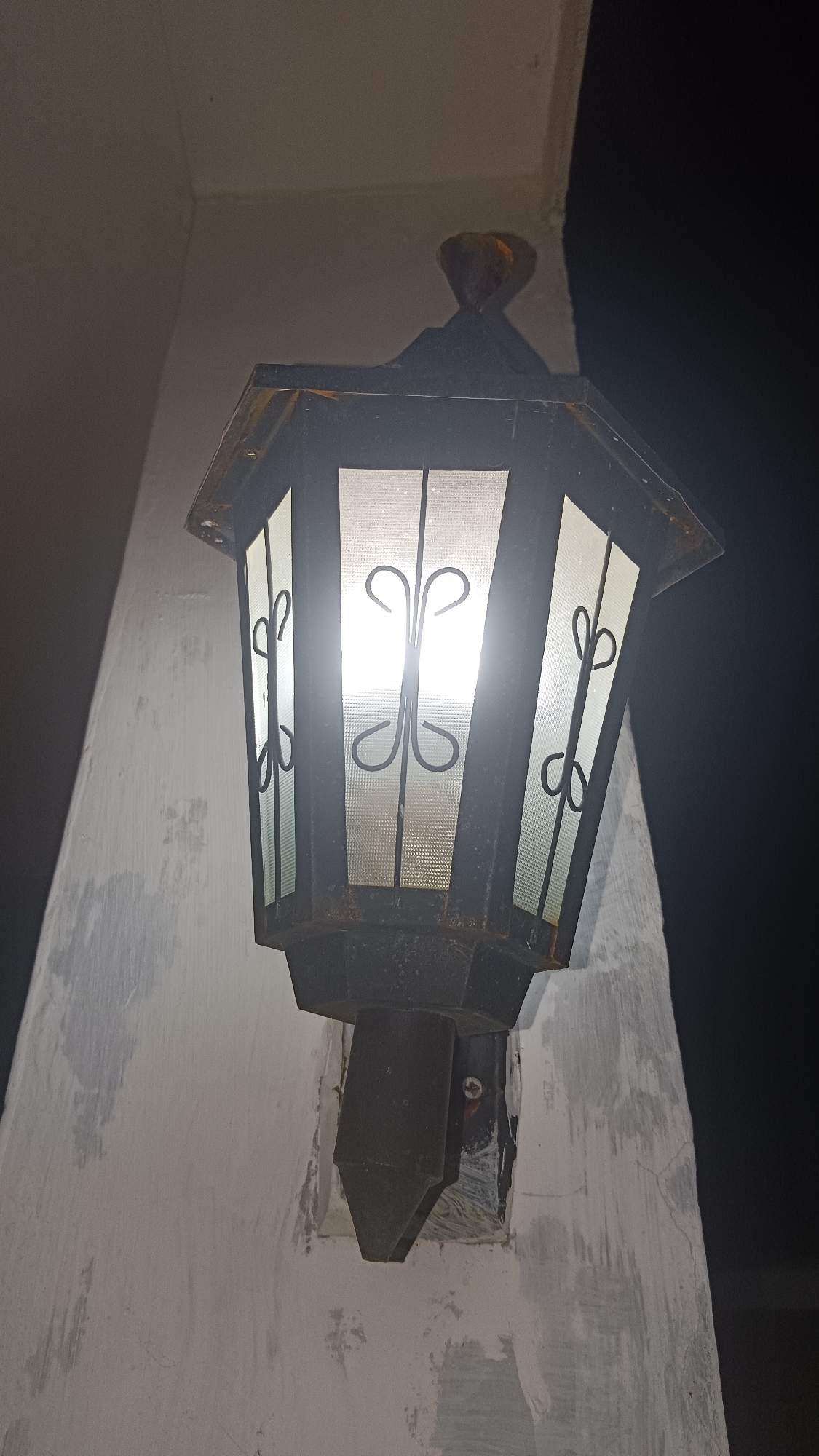 An outdoor, vintage lantern with decorative black metal scrollwork on its frosted glass panels is mounted on a white, textured wall. The lantern is octagonal in shape and features a dark, possibly rusty, metal frame. The light source inside is illuminated, casting a bright glow through the panels. The wall appears weathered, with patches of discoloration and some visible cracks. The background is dark, suggesting nighttime. The scene is set on a wall in Kemang, Indonesia.