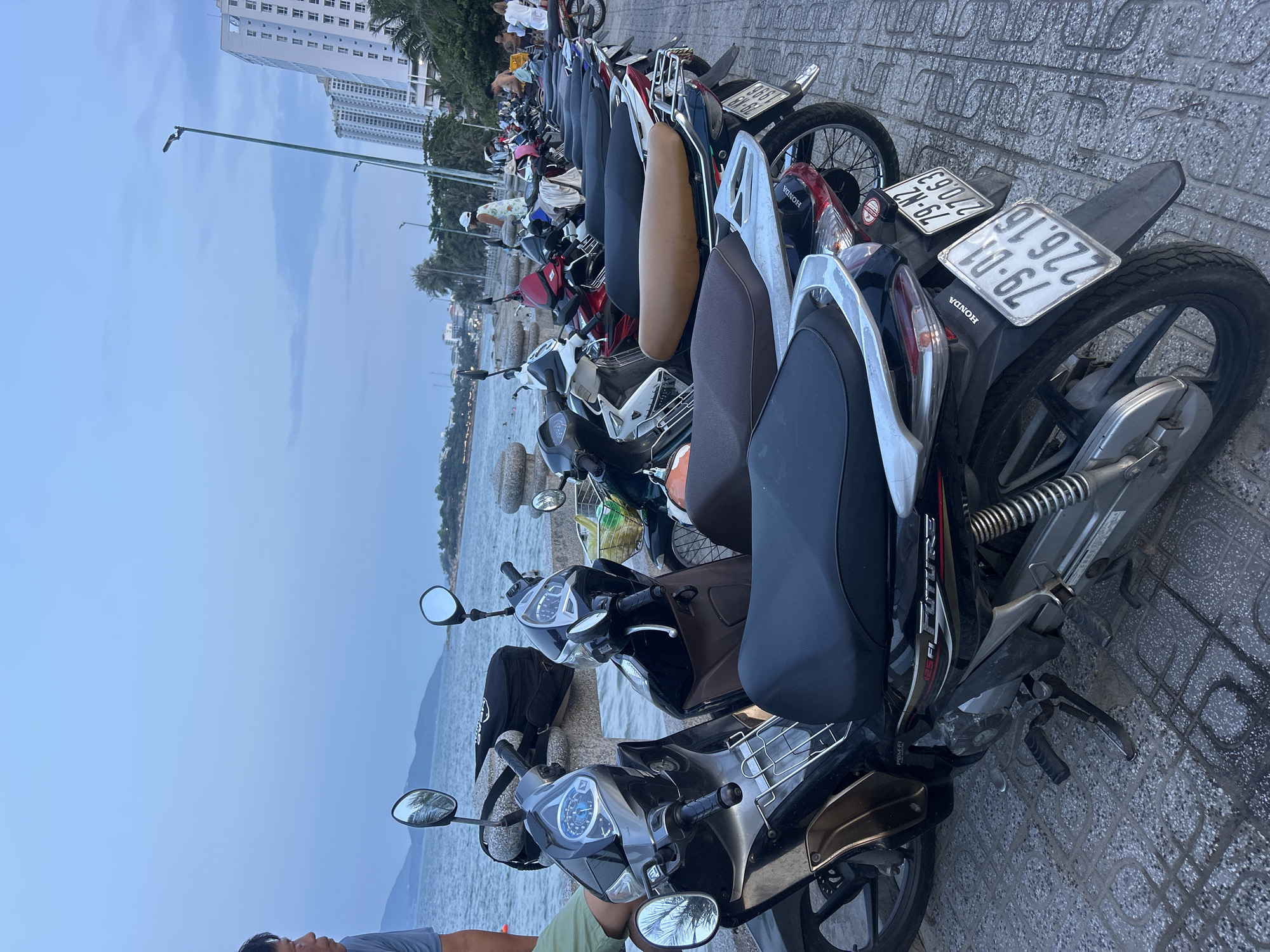 The image shows a row of parked motorbikes on a paved sidewalk in Bac Nha Trang Ward, Vietnam. In the background, there is a body of water, likely the sea, with a coastline and buildings visible in the distance. The sky appears overcast. People can be seen walking along the sidewalk in the distance. The motorbikes are of various makes and models, with license plates visible on some. The paving stones have a grey, patterned texture.