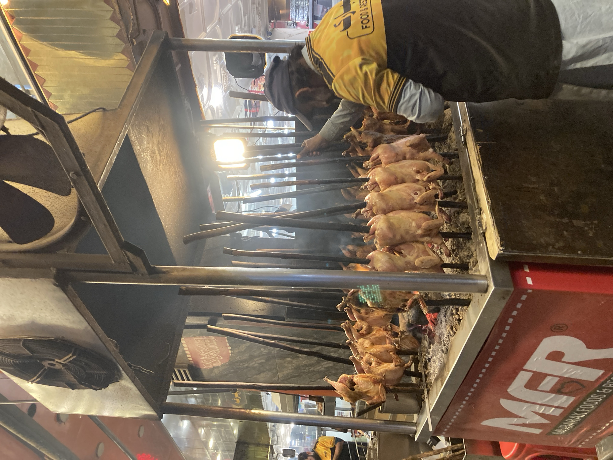 The image shows a street food vendor in Quetta City Tehsil, Pakistan, grilling chickens on skewers over an open fire. The chickens are arranged vertically, with the lower ones more cooked than the upper ones. The vendor, wearing a yellow and black jacket, is attending to the grilling process. The food stall is labeled "MFR Mubarak Foods & Restaurants." The surrounding area is a bustling street with other vendors and activity.