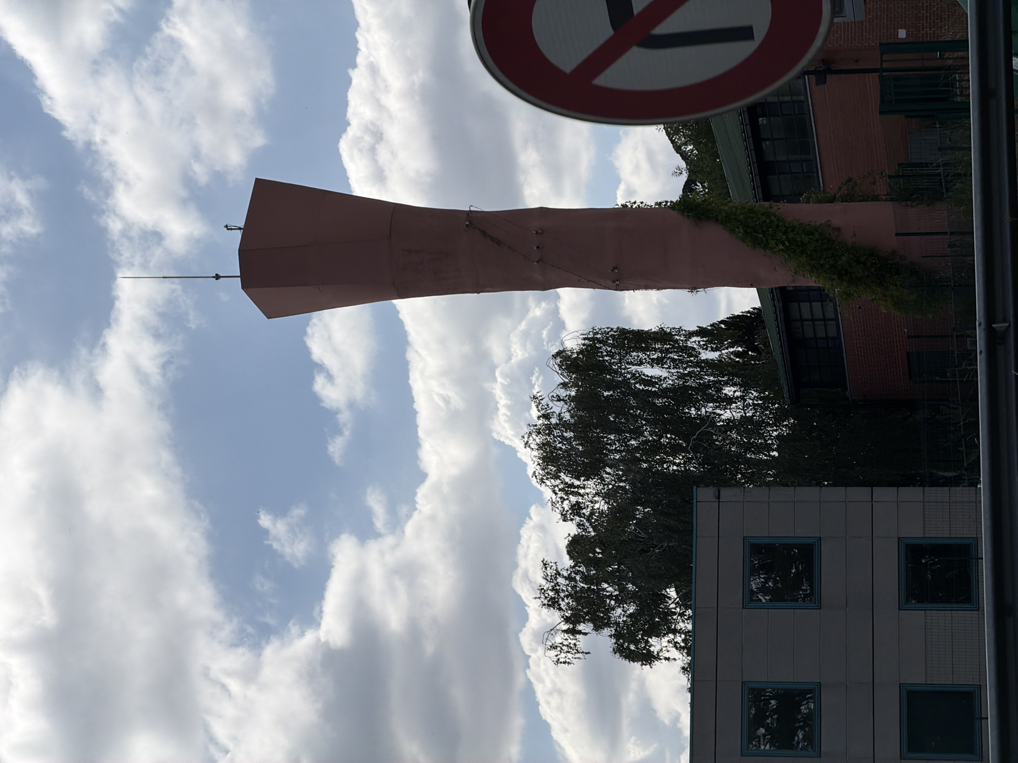 The image shows a tall, conical, pink structure, likely a water tower, in the city of Évry-Courcouronnes, France. The structure has a lightning rod at its apex and is partially covered in ivy. To the left of the water tower is a modern, grey building with several square windows. Behind the water tower and to its right is a red brick building with a green roof. A traffic sign, indicating a prohibition of turning, is visible in the foreground on the right side of the image. The sky is cloudy, with patches of blue visible. The overall scene appears to be an industrial or urban area. There are no people visible in the image.