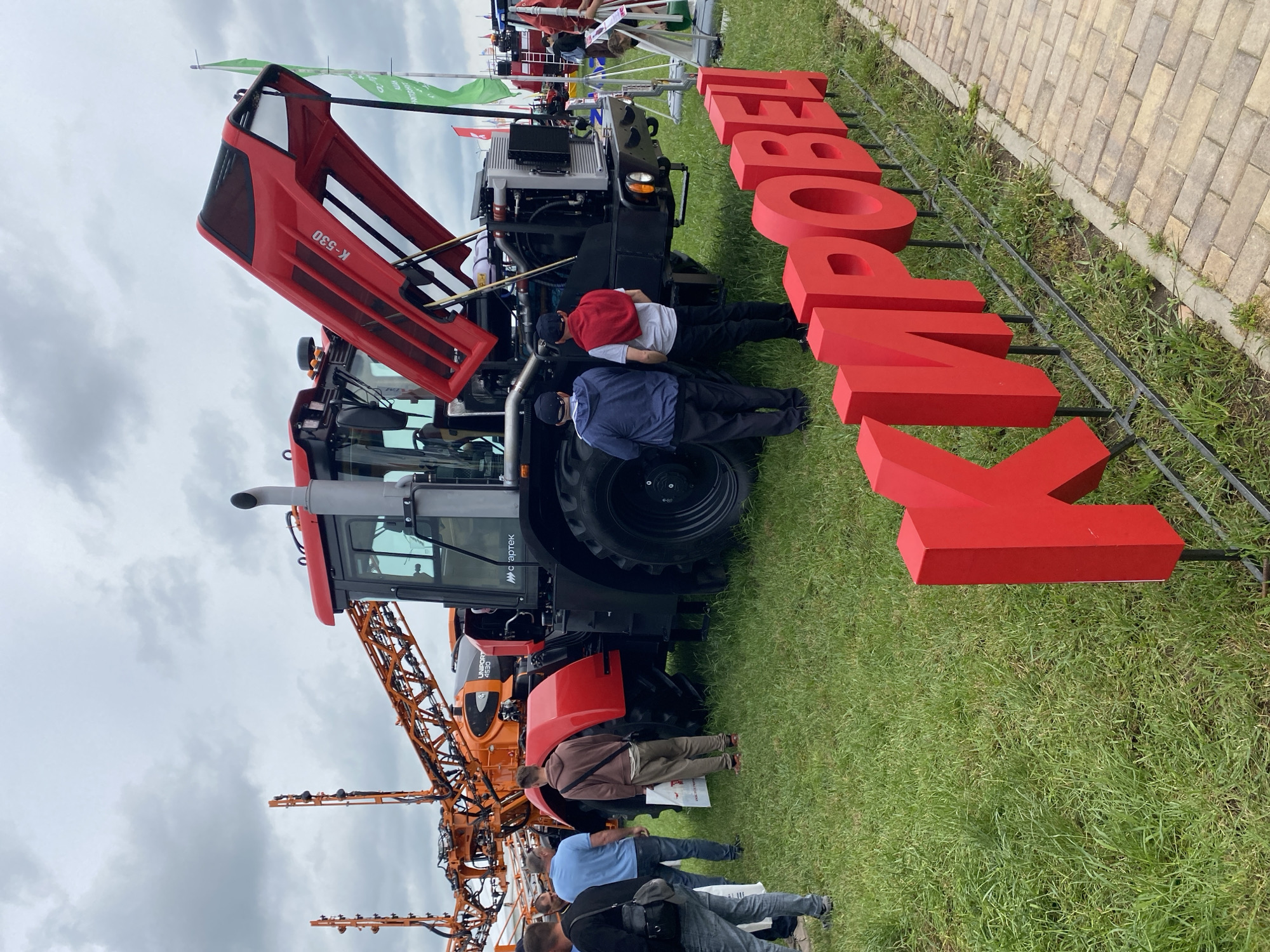 The scene takes place outdoors on a grassy area adjacent to a brick-paved walkway, under a cloudy sky. The primary focus is a large red tractor with its hood raised, revealing its engine. Lettering "KIMPOBEL" in large red, three-dimensional letters stands to the side of the tractor, supported by a black metal frame. A second agricultural vehicle, possibly a Uniroot 2530, with an orange frame and spray booms, is visible behind the tractor. Several people are present: some are looking at the tractor’s engine, while others stand further away, observing the equipment. The location is specified as Voronezhskaya, Russia. The people appear to be interacting with the agricultural machinery, potentially examining or learning about its features.