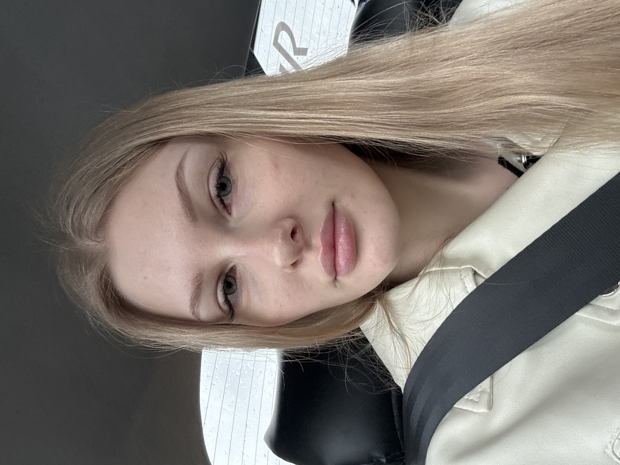 A young woman with long, straight blonde hair, light skin, and green eyes is in a car, wearing a seatbelt and a light-colored jacket. The interior of the car is visible. The letter "R" is visible in the background above her head. There is no evidence to determine that the location is Perm, Russia.