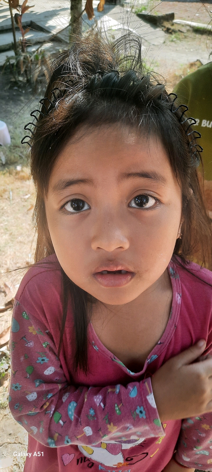 A close-up, low-angle shot focuses on the face of a young girl in Ploso, Indonesia. She has dark hair with strands sticking up, held back by a black, wavy hair accessory. Her eyes are large and dark, with reflections of lights visible in them. Her mouth is slightly open, revealing missing teeth, and her lips are a soft pink. She is wearing a long-sleeved pink shirt with a pattern of mushrooms, flowers, and small pots. One of her arms is crossed over her chest.
The background is softly blurred, suggesting an outdoor setting. Sunlight appears to be present, indicated by the bright lighting and shadows in the background. To the right of the girl's head, part of a green garment and some text are visible, though it is mostly out of focus and obscured. The bottom left corner of the image displays the text "Galaxy A51". The scene appears to be informal and candid, capturing a moment with the child.