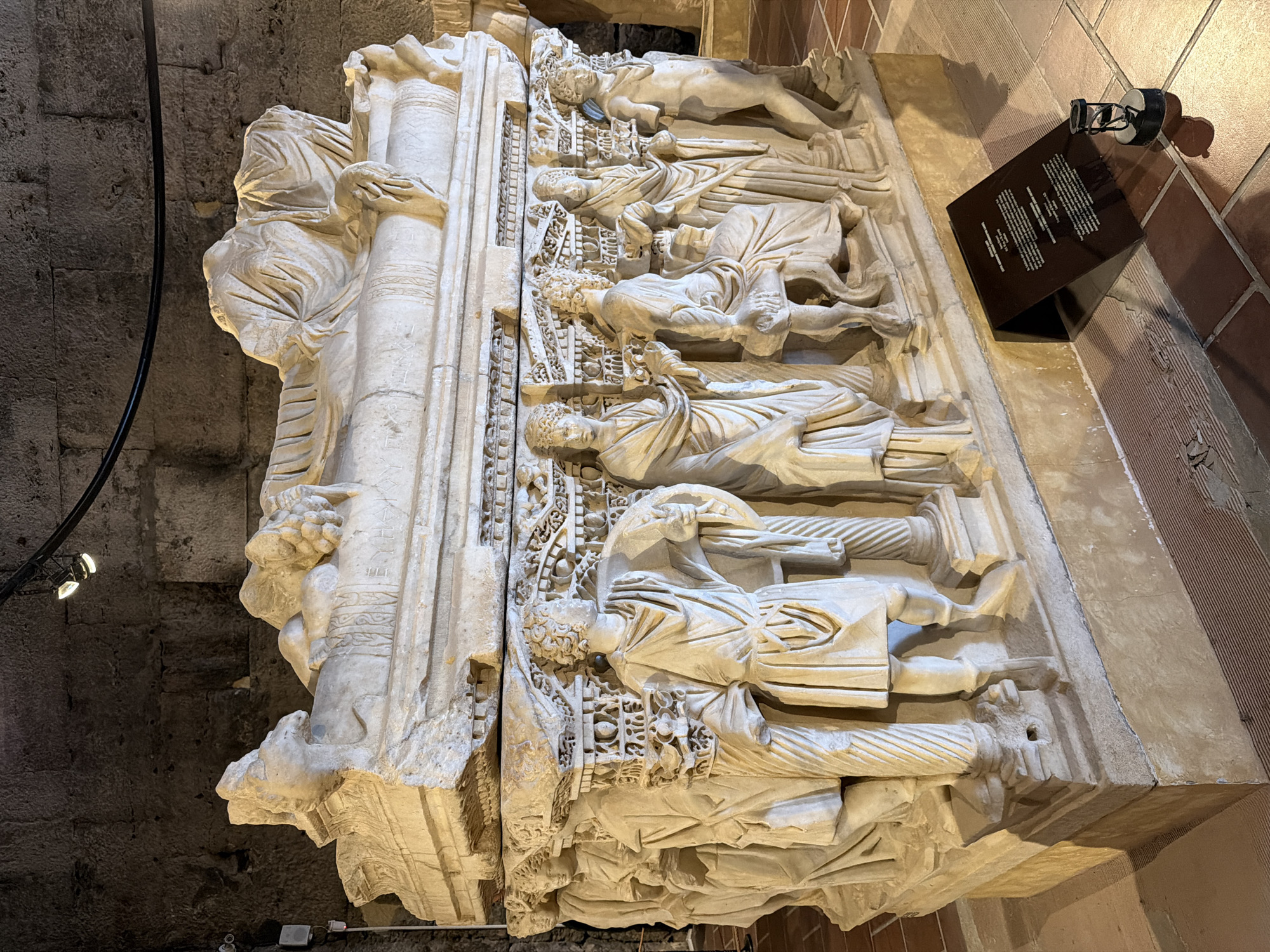 The image shows a large, weathered white marble sarcophagus with intricate bas-relief carvings, positioned inside a stone structure with brickwork visible on the right side. The carvings depict human figures in classical robes, arranged in tiers. Above and to the right of the sarcophagus, there is a dark, angled information plaque with printed text. The sarcophagus is displayed under artificial lighting, with light fixtures visible in the upper left corner. Based on the architectural details and likely historical context, the sarcophagus could potentially be located in a museum or historical site in Pamukkale, Turkey, given the presence of ancient ruins in that region.