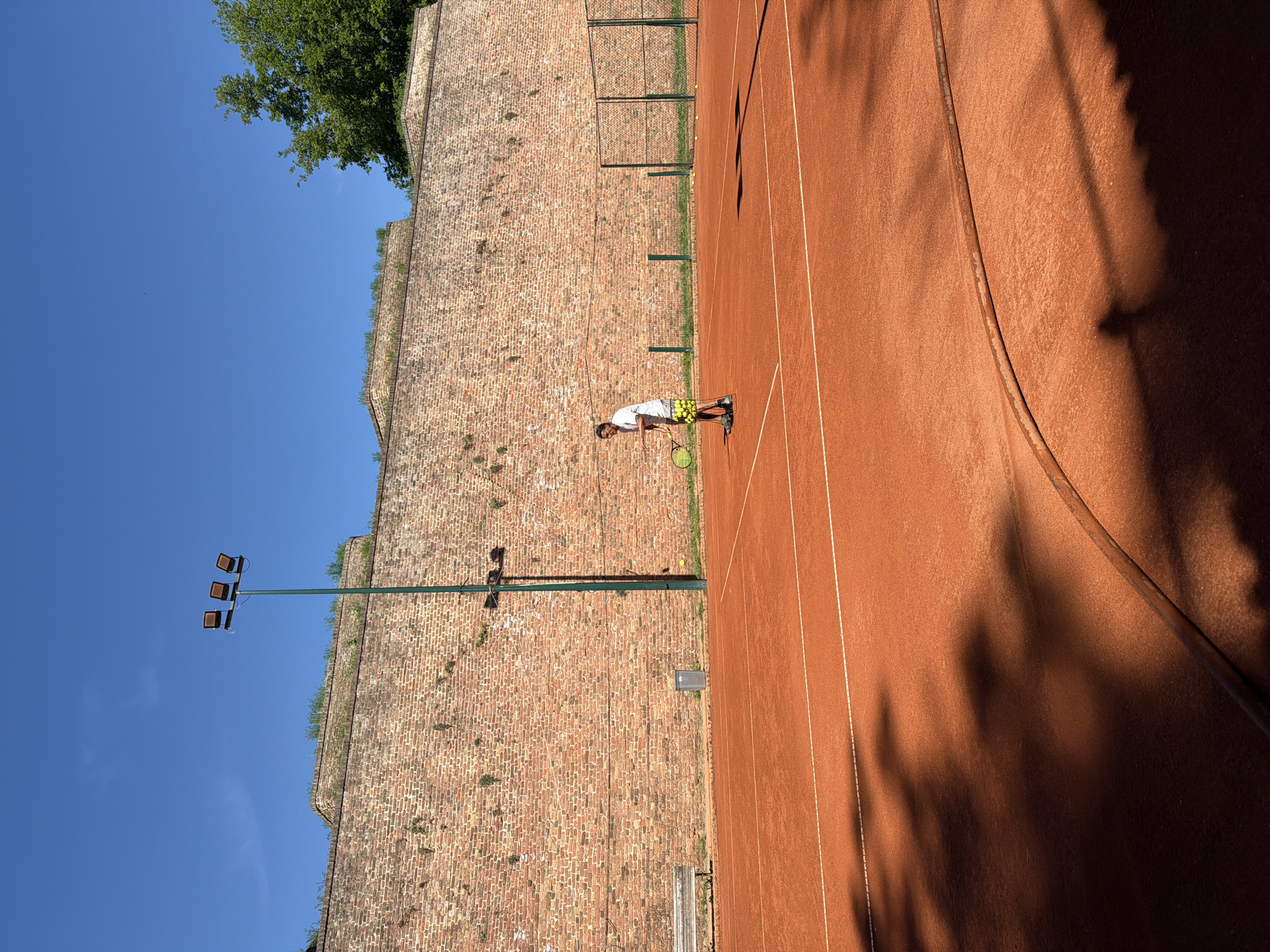 The image depicts a tennis court adjacent to a high brick wall, under a clear blue sky. The court's surface is red clay. A man in a white shirt and patterned shorts stands on the court, holding a tennis racket. Several tennis balls are scattered near the back of the court. A tall light pole with three lamps is situated near the wall. Vegetation grows on the top of the wall. The location is within the Kalemegdan Fortress in Belgrade, Serbia, as indicated by the fortress wall construction. The presence of a clay tennis court is significant.