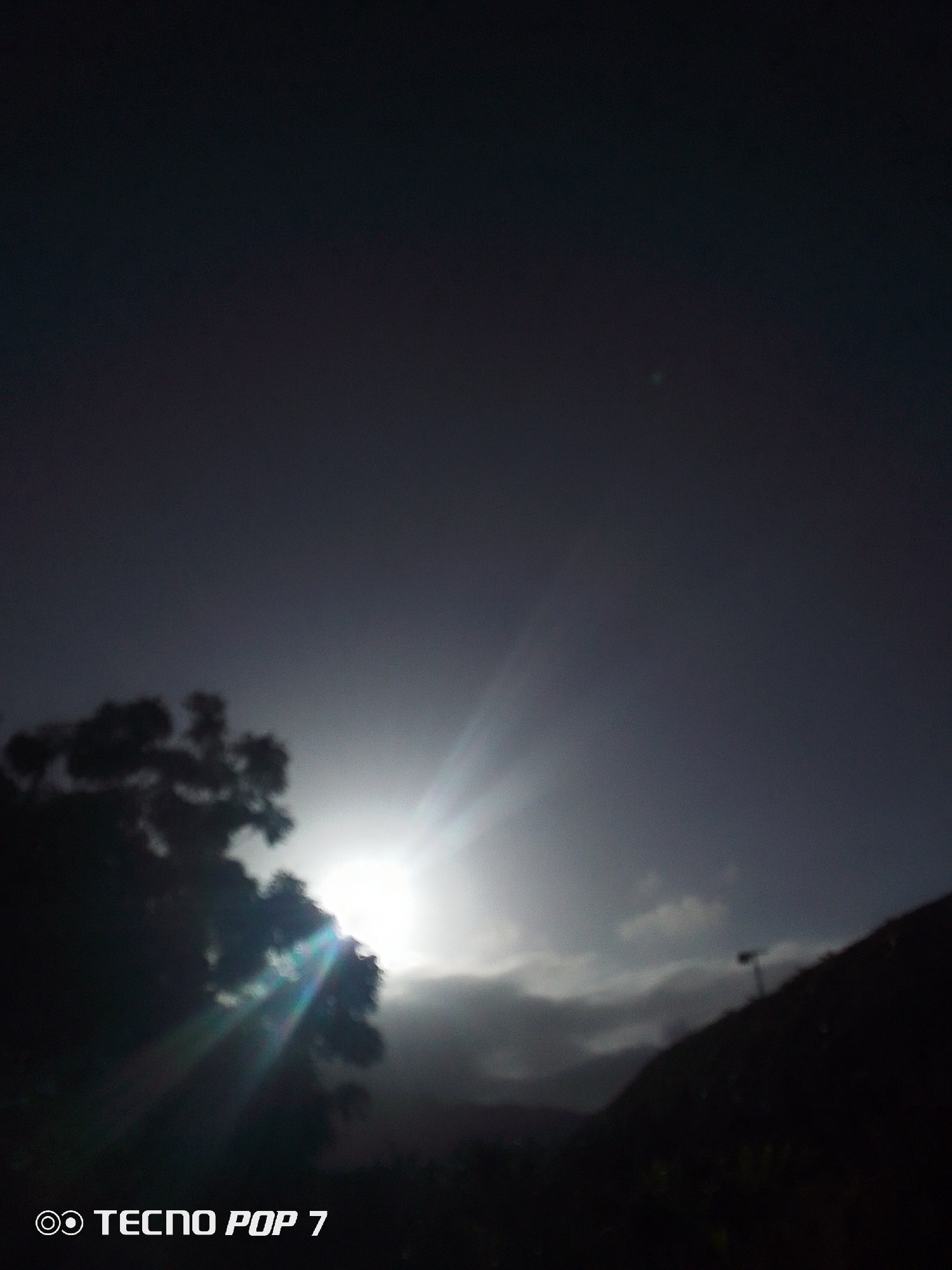 A low-angle photograph depicts a landscape in possible Pedro González, Venezuela, captured during twilight or early morning. The focal point is a radiant light source, likely the sun, obscured by either a tree or heavy cloud cover. The light rays emanate dramatically. To the left, the silhouette of a tree is prominent, while a dark, elevated landmass is on the right. A layer of clouds lies above the landmass. The top half of the image is a dark gray sky. The lower left corner of the image has the text "TECNO POP 7," indicating the camera used.