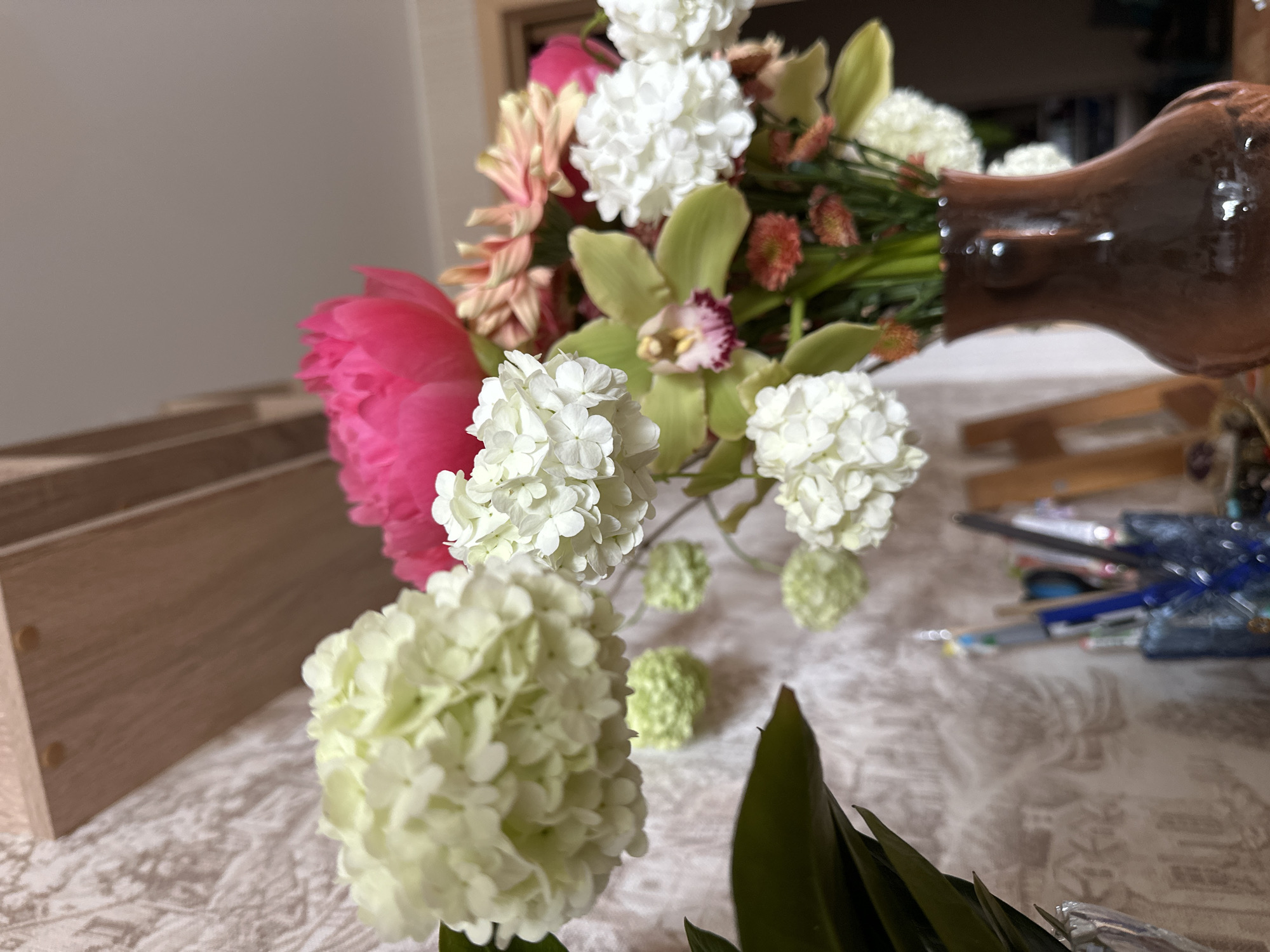 The image presents a close-up view of a floral arrangement, possibly indoors. A bouquet of flowers includes white hydrangeas, a pink peony, light green orchids, and other flowers of various colors and types. The flowers are arranged in a brown ceramic vase. The arrangement sits on a table covered with a patterned tablecloth. To the left is a light-colored wooden box. In the background are stationery items like pens and pencils and other objects on a table. The location, Vladimir, Russia, is not determinable from the visual information provided.