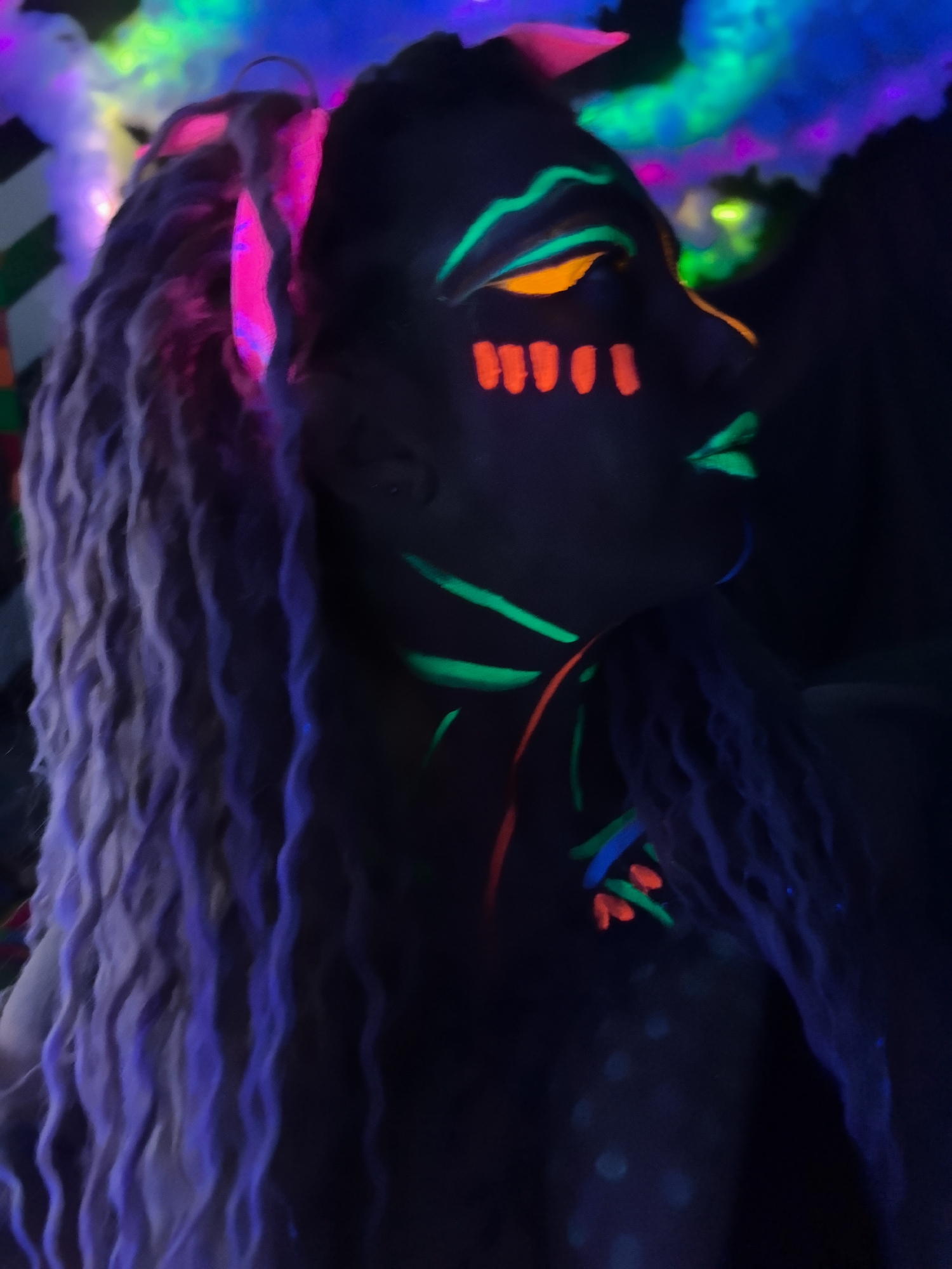 A profile view shows a person with light-colored dreadlocks and neon face paint in a dark environment. The face paint is applied in stripes above the eyes, short vertical lines under the eyes, lines on the neck, and highlights on the lips. The colors of the paint include orange, green, and pink. Some of the dreadlocks are illuminated with a violet glow. The background is dark with indistinct areas of green, blue, and pink light. The location is unknown. The city of Noviy, Russia is not mentioned in the image.