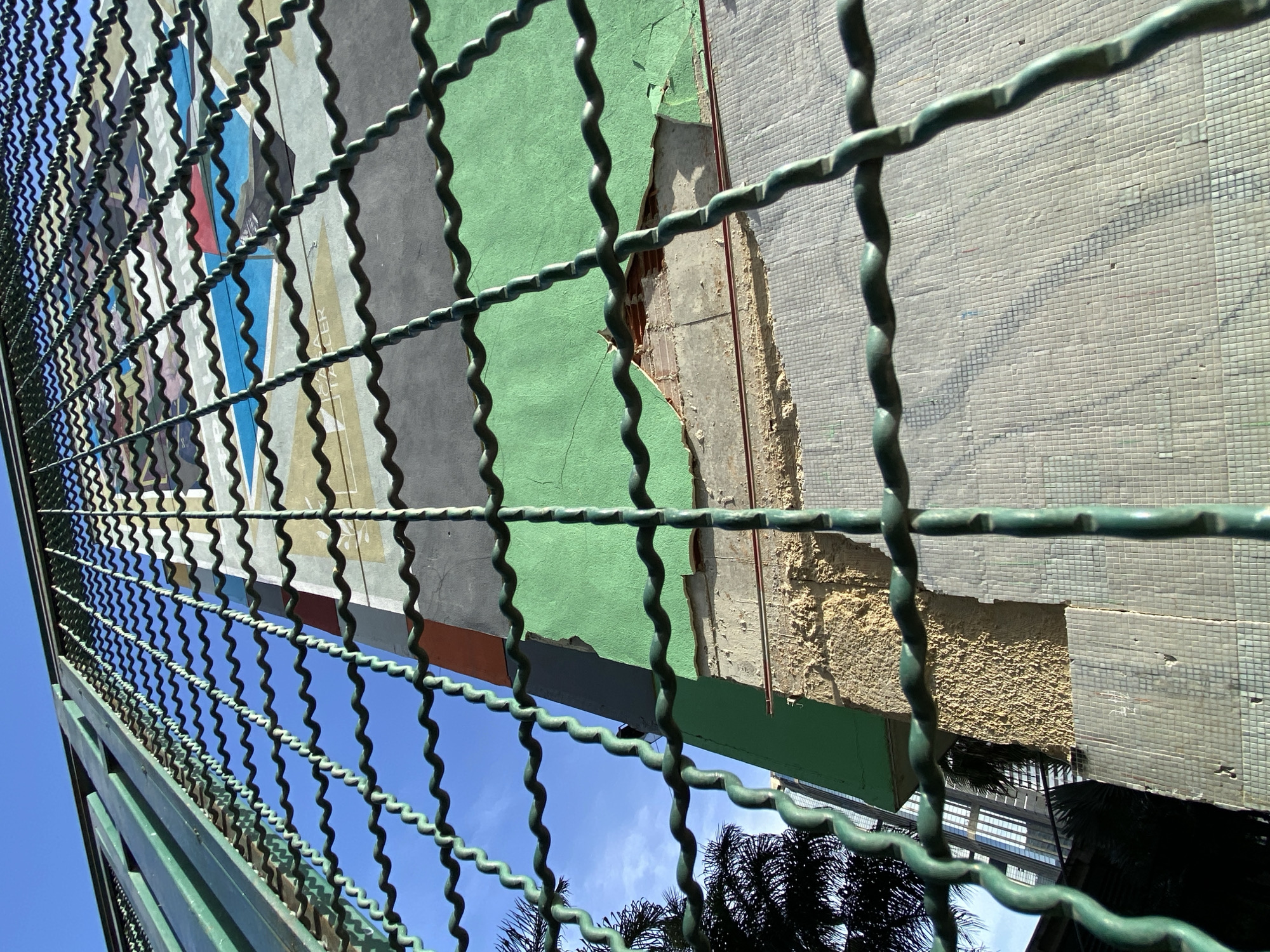 The image shows a close-up view of a building facade, partially obscured by a green metal fence. The building's wall is a patchwork of different materials and colors. In some areas, there is light gray concrete with a mosaic tile finish, which appears worn and incomplete in places. Other sections feature a vibrant green painted surface that is peeling away, revealing the rough concrete underneath. There are also fragments of other colors, including orange and what appears to be a mural with blue, red, and yellow elements. The wall shows signs of damage and disrepair, with large sections of plaster missing, exposing the underlying brickwork.
The perspective is from a low angle, looking up towards the building. The sky is a clear, bright blue, suggesting it is daytime. The sun is casting shadows, indicating it is likely midday. In the lower part of the image, the tops of dark green palm fronds are visible, suggesting a tropical or subtropical climate. In the distance, through the fence and the gaps in the building, a modern glass-fronted building can be seen, hinting at an urban environment. The overall impression is one of urban decay juxtaposed with nature and modern architecture. There is no discernible activity or event taking place. No text is visible. Given the location context of Caracas, Venezuela, the scene likely depicts a building in an urban area undergoing renovation or suffering from neglect.