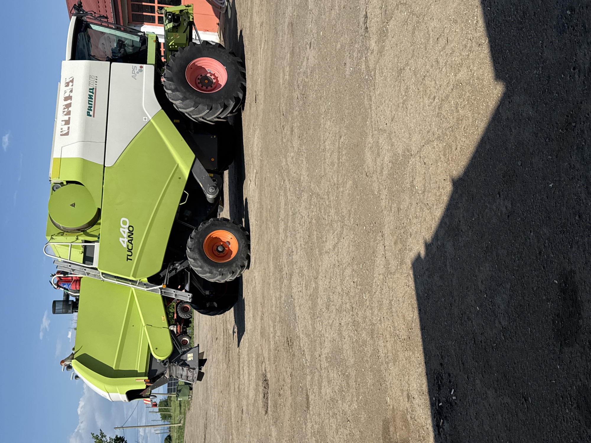 The image shows a green and white Claas Tucano 440 combine harvester parked on a concrete surface. The harvester is oriented from left to right in the frame. The tires are orange and red. The background includes a partial view of a red-roofed building and a patch of blue sky. The right side of the image is dominated by a large shadow on the concrete. The location is Sarnevo, Bulgaria.