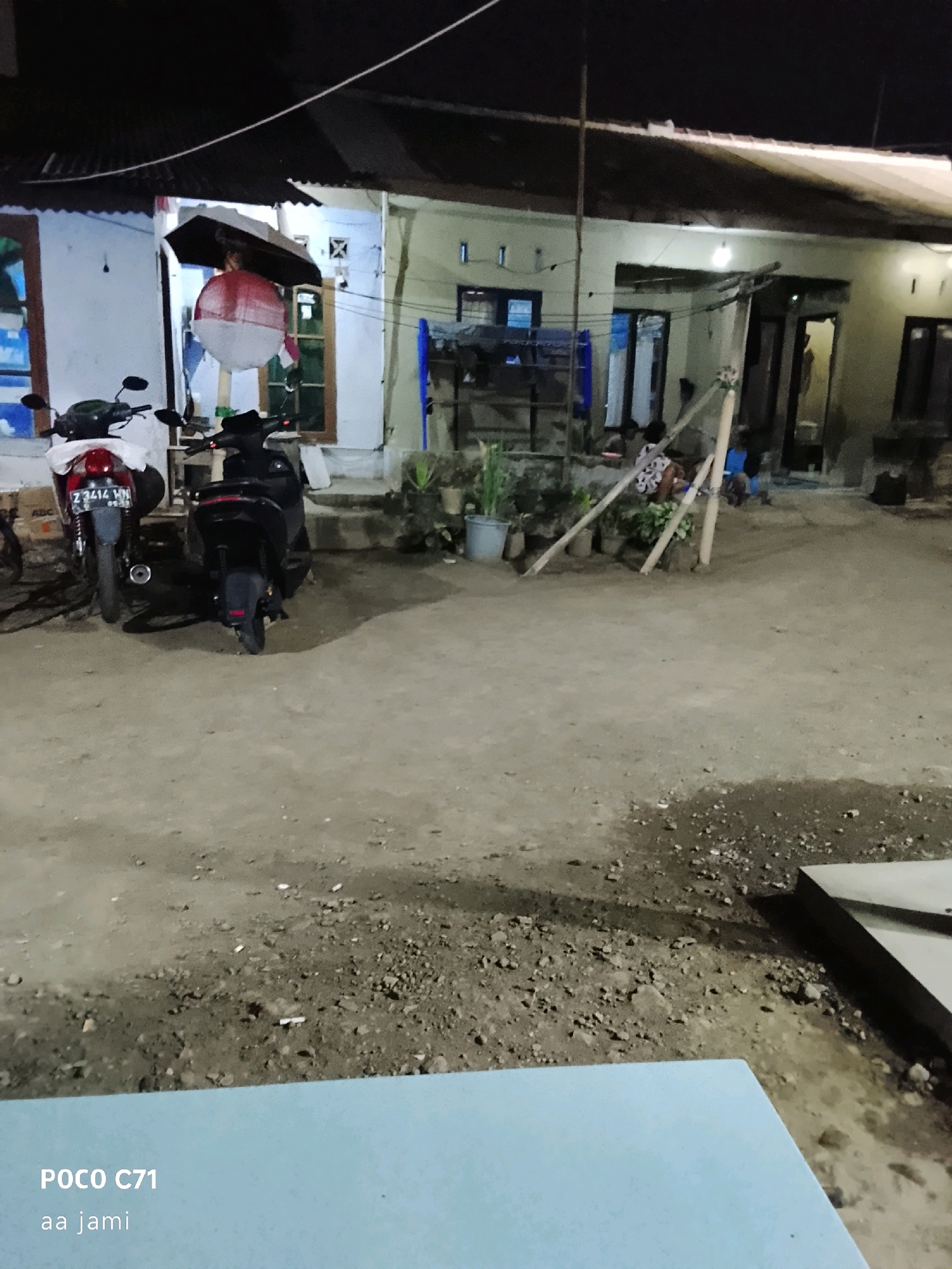 The image shows a dark, outdoor scene at night, likely in Tasikmalaya, Indonesia, as indicated by the text overlay "POCO C71 aa jami". The foreground is dominated by a light blue surface. In the mid-ground, there is a dirt or gravel ground with scattered debris, and two motorcycles are parked. One motorcycle, on the left, is partially covered with a white cloth and has a license plate that reads "Z 3414 VN". The other motorcycle, on the right, is black. Beyond the motorcycles, there is a building with a corrugated roof. Under an umbrella, a large, red and white spherical object, possibly a lantern or decoration, is suspended. The building appears to be a modest residential structure with several doorways and windows. In the background, several people are visible, some sitting near the building. The overall impression is of a residential area at night, possibly during a local event or festival.