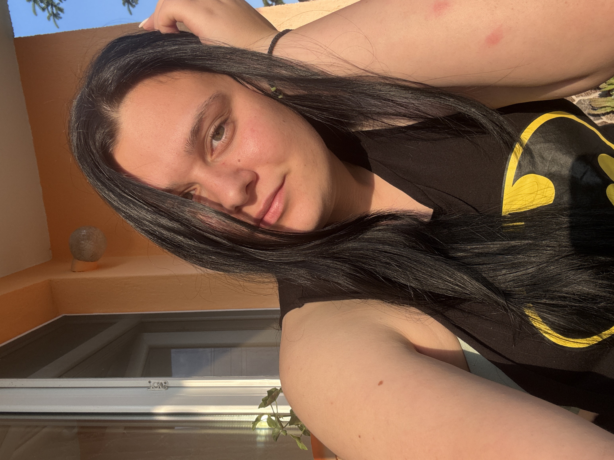 A young adult with long, dark hair and light to medium skin tone is depicted in a close-up, chest-up outdoor portrait taken in Rogosh, Bulgaria. The individual is looking directly at the camera with a neutral to slight smile, their head tilted slightly. Long, dark hair is parted on the viewer's left, with some strands falling over the right side of the face. The person is wearing a black sleeveless top or tank top with a yellow Batman logo visible on the chest.
The individual's right arm (viewer's left) is partially visible, showing a small dark mole on the upper arm. The left arm (viewer's right) is bent, with the hand positioned behind or on the back of the head. A black bracelet is present on the left wrist. Two distinct reddish marks are visible on the upper left arm. A small green earring or bead is visible on the left earlobe.
The background shows an outdoor setting. To the viewer's left, a white-framed window or door with glass panes is visible, reflecting an interior space or another window. A small green potted plant is situated near the window frame. To the viewer's right, an orange or terra cotta colored wall is visible, with a spherical outdoor light fixture mounted on it. Above the wall, a clear blue sky and green foliage from a tree are discernible. Strong natural sunlight illuminates the scene, casting highlights on the individual's hair and skin.