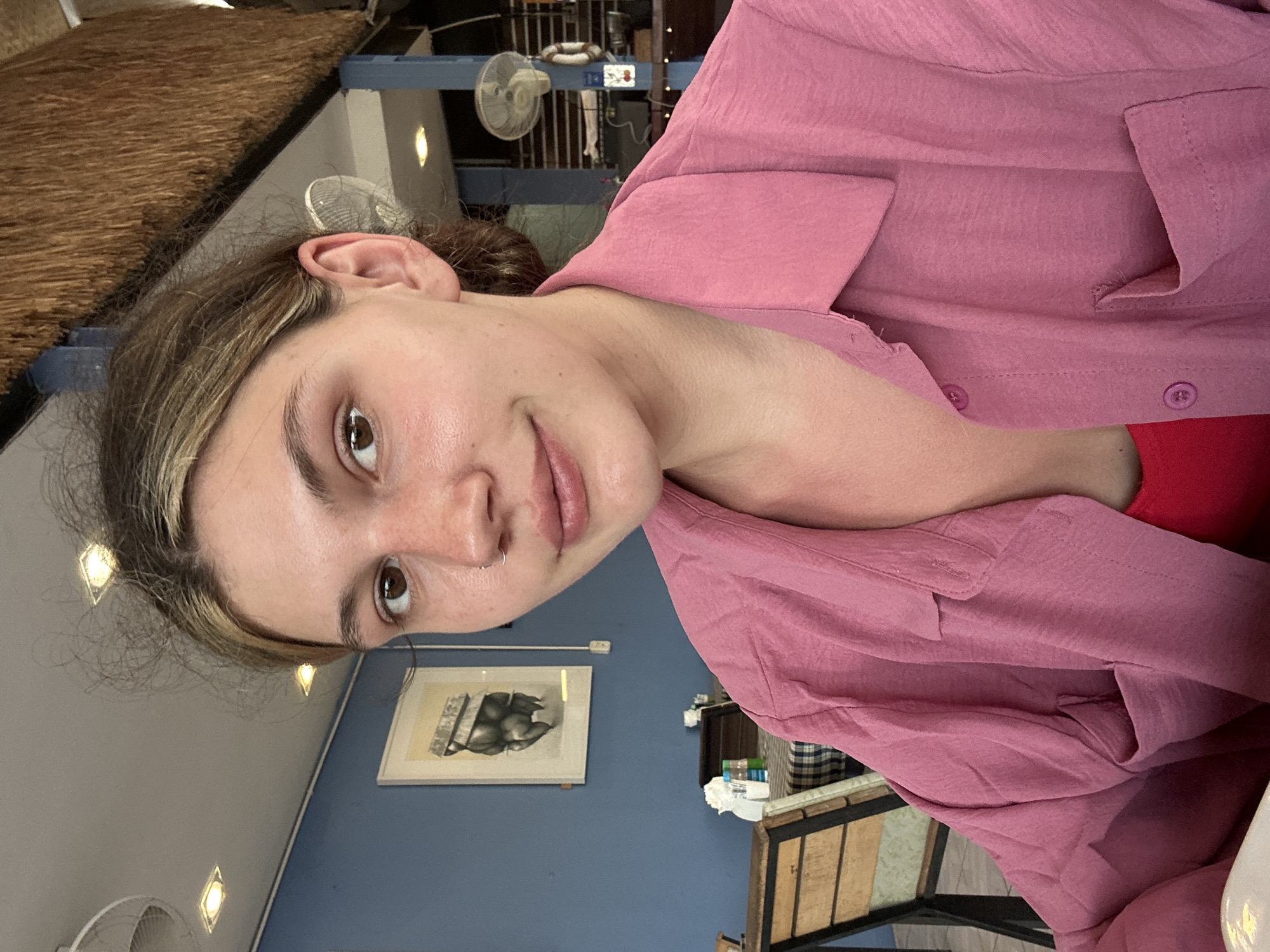 The image shows a young woman in a pink button-down shirt. She has brown eyes, a nose ring, and her hair is pulled back. The background features a blue wall with a framed print of an animal, possibly an elephant or bear. There's a ceiling with recessed lighting and a thatched roof section visible. A fan is attached to a blue structural support beam. The objects present and building style suggest the location is Baan Chaweng, Thailand.
