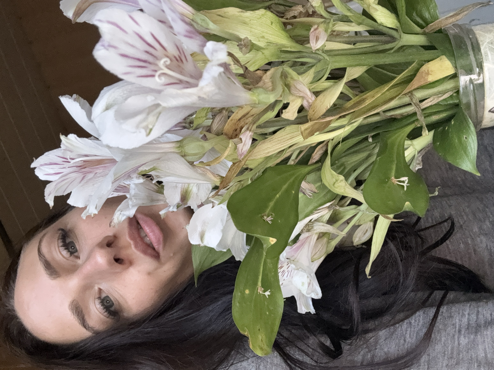 This image depicts an indoor scene in Saint Petersburg, Russia. The primary subject is an adult female, positioned in the upper left quadrant of the frame, looking directly towards the viewer. She has long, dark brown hair, fair skin, and light-colored eyes with visible eyelashes, possibly enhanced with mascara. Her lips are full and slightly parted. She is wearing a grey garment, likely a shirt or sweater, visible on her shoulder and lower left side.
A bouquet of white flowers with prominent pink-to-purple vertical stripes, characteristic of Alstroemeria (Peruvian Lily), occupies the central and right portions of the image. Several flower heads are open, displaying their distinctive patterns, while others are less open or in bud. Some of the petals and numerous leaves exhibit signs of senescence, with browning and desiccation evident. The stems of the flowers are gathered in what appears to be a clear glass jar or vase, visible at the bottom right. The background behind the woman in the upper left is a solid, dark brown surface. The background behind the flowers and the lower parts of the image is a lighter, uniform grey, consistent with the color of the woman's clothing. The lighting appears to be even, without harsh shadows.