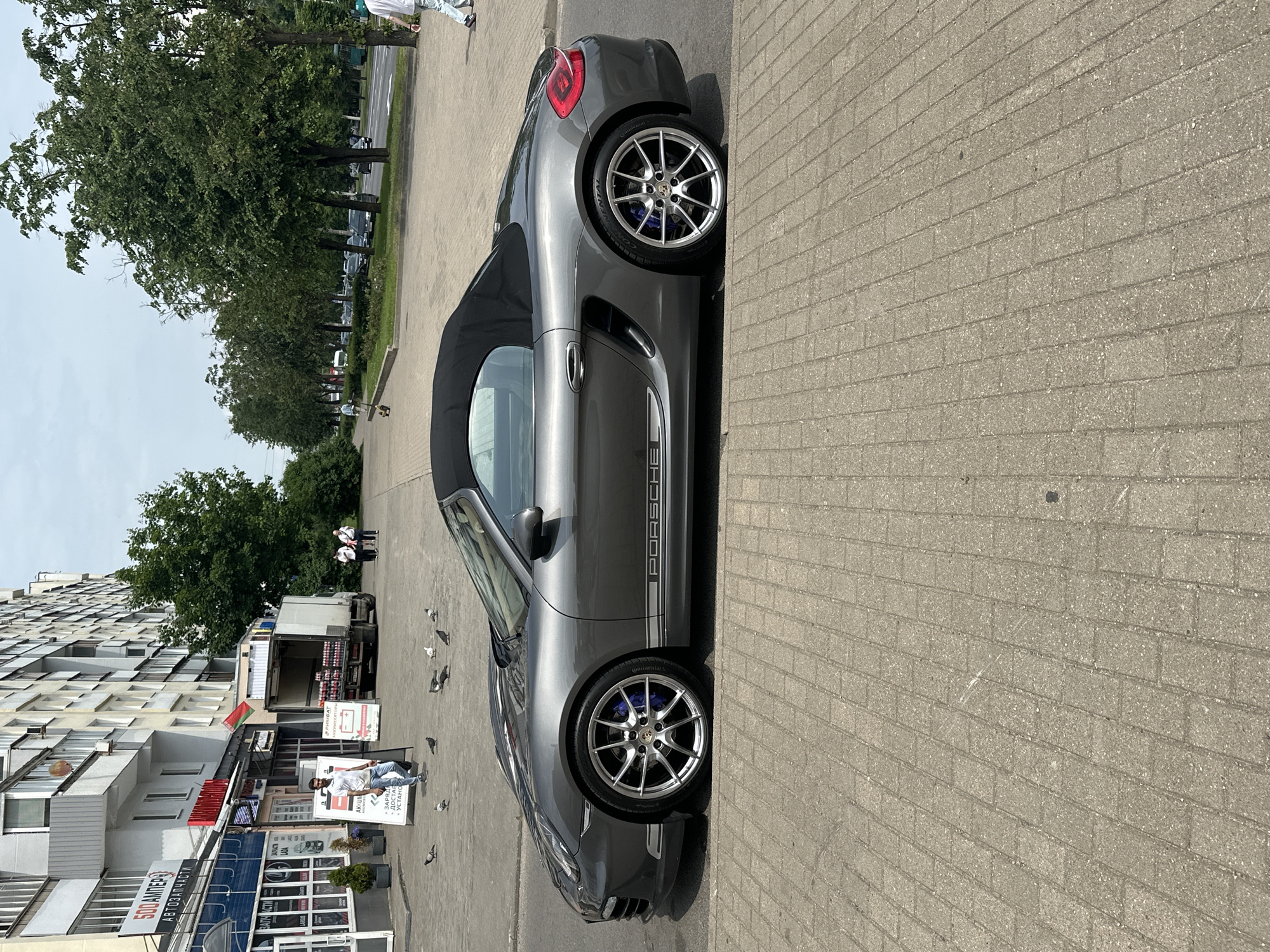 A grey Porsche Boxster convertible is parked on a brick sidewalk in Minsk, Belarus. The car is facing right, with its black soft top up. The front and rear wheels have silver rims with blue brake calipers. The word "PORSCHE" is visible in white lettering on the side of the car. In the background, there is a multi-story building with shops on the ground floor, a delivery truck, trees, and a street with traffic. A man in a white t-shirt and blue jeans is walking away from the camera, and several pigeons are on the sidewalk near the car. The sky is overcast.