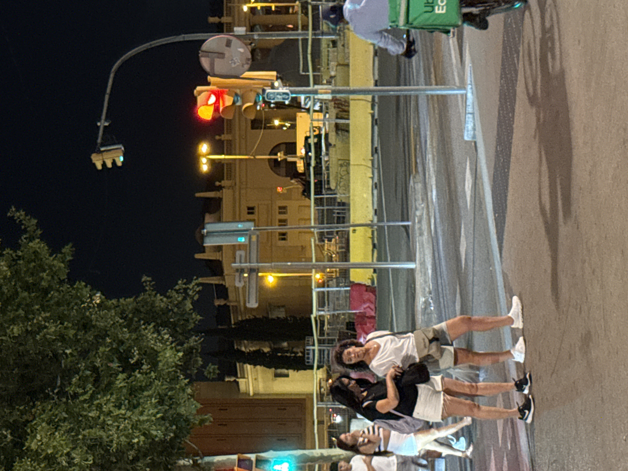 This image captures a street scene at night in Barcelona, Spain, likely near a construction zone. In the foreground, two women stand on a crosswalk. The woman on the left wears a black t-shirt and white shorts, with black and white sneakers, and carries a dark shoulder bag. The woman on the right is dressed in a white t-shirt and light-colored shorts, with white sneakers, and also carries a shoulder bag. Behind them, other pedestrians are visible, including a man in a white t-shirt and a woman in a striped top and white shorts.
To the right, a person on a bicycle with a bright green "Uber Eats" delivery bag is moving away from the camera. A traffic light with a red signal is prominent in the middle ground, positioned above yellow construction barriers. The background reveals a large, light-colored building with arched entrances and windows, possibly a public or commercial structure. Streetlights illuminate the scene, and a large tree with dense foliage is visible on the left side of the image. The overall atmosphere suggests a busy urban environment at night, with ongoing construction affecting the public space.