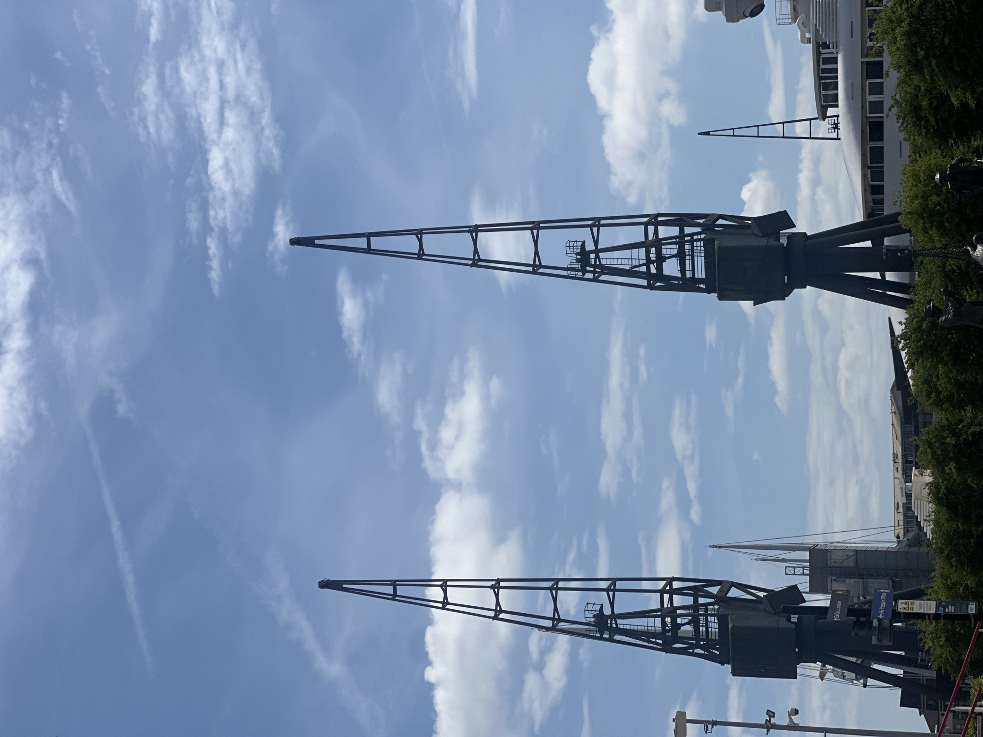 The image depicts the preserved historic dockyard cranes in the former port area of London, United Kingdom, under a partially cloudy sky. Two prominent, dark-colored cranes dominate the foreground, their latticework structures pointing upwards. In the background are other cranes, part of the dockyard complex and a large white building, possibly a modern structure or ship. Trees and other urban elements are visible to the side of the frame, indicating the location is now integrated within the city. The angle is looking upwards, emphasizing the height of the cranes.