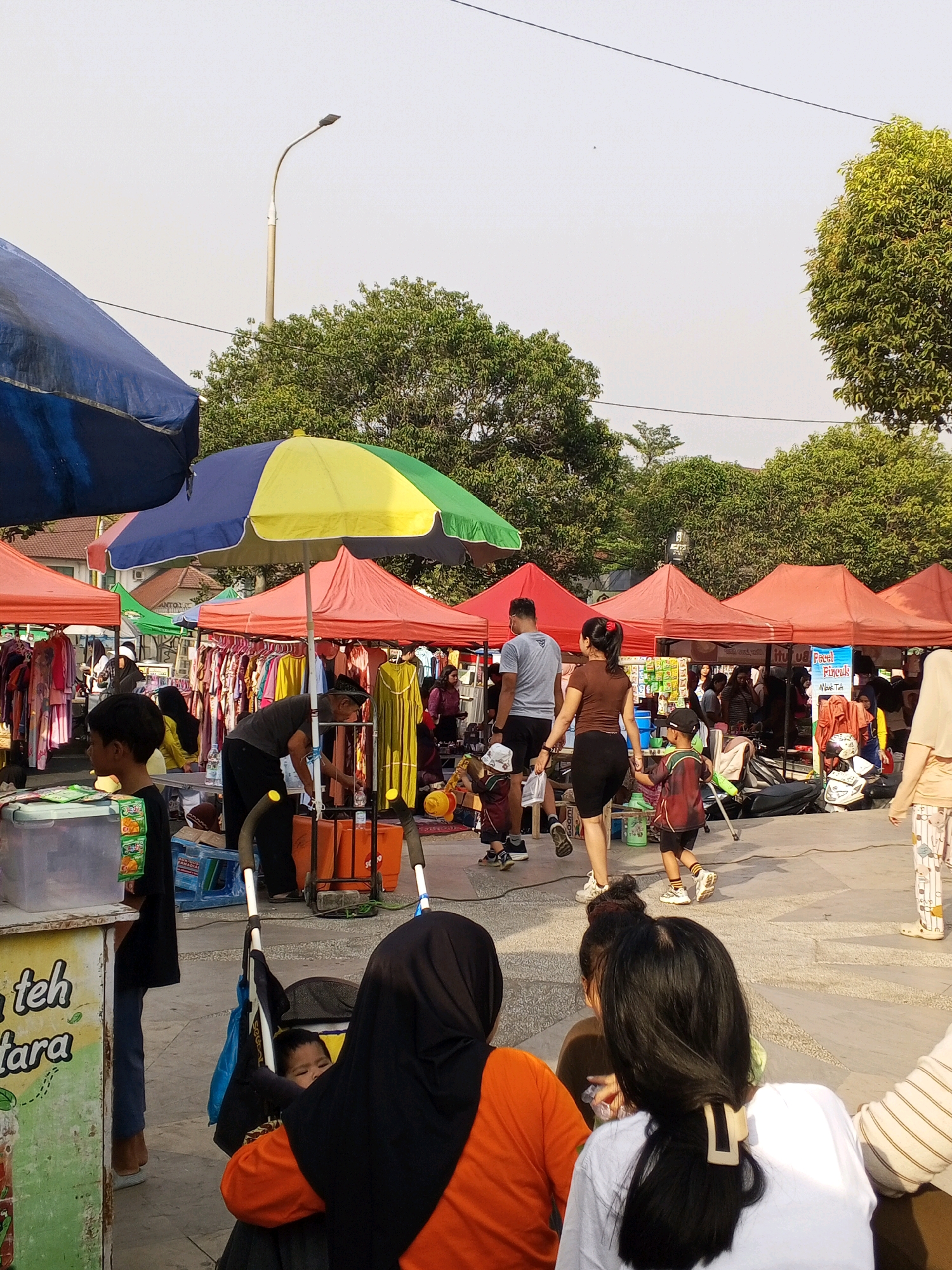 The image depicts an outdoor market scene, likely in Serang, Indonesia. The environment is characterized by numerous stalls covered with red, yellow, and green awnings. Clothing racks filled with garments are visible behind some of the stalls. Several people are browsing or walking through the market. In the foreground, a young boy in a black t-shirt is looking to the left. To his right, a man wearing a black cap is attending to a stall. Further back, a couple with a child is walking away from the camera. A stroller with a baby inside is also present. The ground is paved, and trees are visible in the background, suggesting an urban park or open public space. A street lamp is visible in the upper left portion of the image. The overall atmosphere suggests a busy daytime market environment.