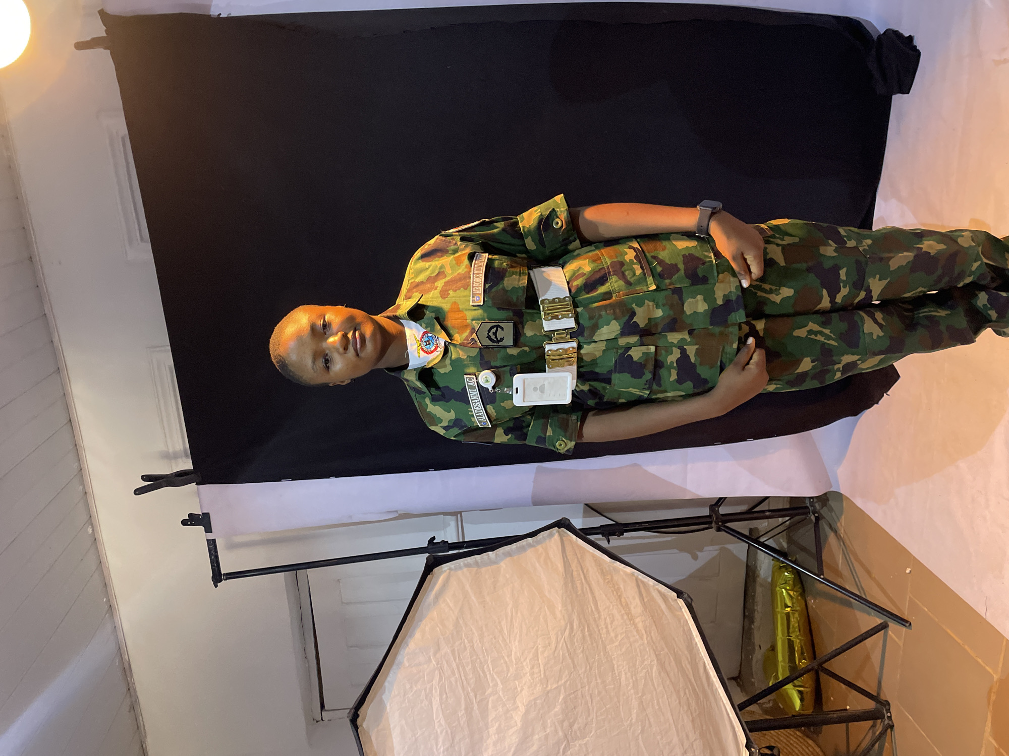 The image shows a person wearing a military camouflage uniform standing in front of a black and white backdrop. The person has short hair, a wristwatch on their left wrist, and several patches and tags on their uniform. The uniform includes a white belt with metal buckles and patches with names or designations. A white ID badge is attached to the front of the uniform. The backdrop is supported by a stand with clips visible at the top left of the frame. A light source with a white diffuser is visible in the foreground. The location is a room with white walls and ceiling. There is no information in the image to suggest the location is in Jos, Nigeria.
