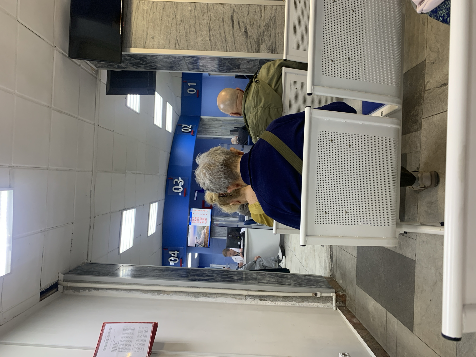 The image depicts an indoor waiting area. Several individuals are sitting on white, perforated chairs, facing away from the camera. Behind them, blue panels with numbers "01," "02," "03," and "04" are visible, suggesting a queuing system. A television screen displays information, possibly related to the services offered. A person wearing a white coat is visible behind a counter. The setting suggests a government office or a public service area. The location is in an unidentified city.