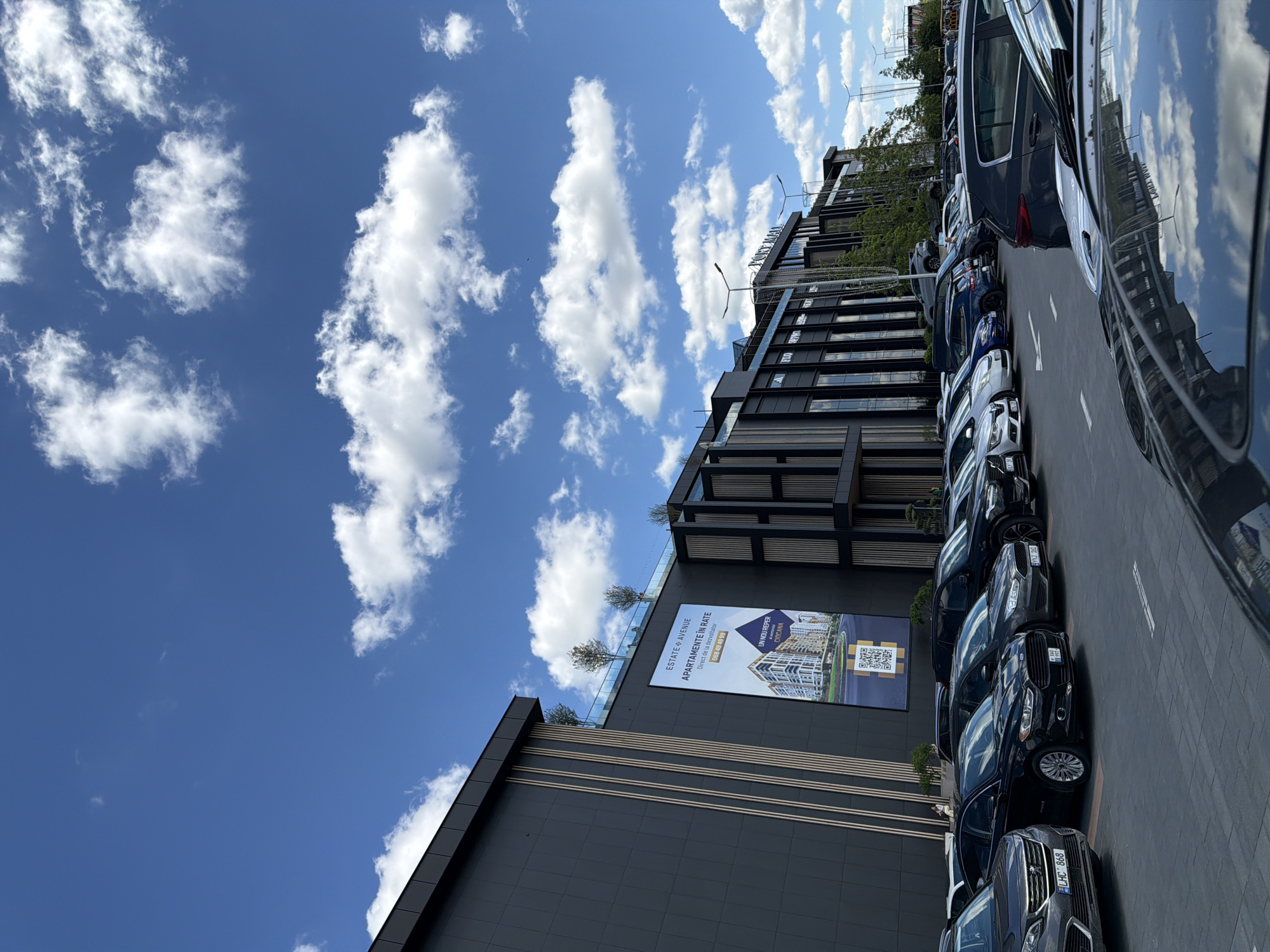 The image depicts a modern commercial complex with a parking lot in front, located in Stăuceni, Moldova. The complex features a dark facade with large windows and architectural elements like vertical wood-like panels and a glass railing on the upper level. A large banner advertisement for "ESTATE & AVENUE APARTAMENTE IN RATE" is prominently displayed on the building's facade.
The parking lot is filled with a variety of cars, primarily dark-colored sedans and SUVs, parked in marked spaces. The ground of the parking lot appears to be paved with dark tiles. In the background, trees and streetlights are visible.
The sky is a bright blue with scattered white cumulus clouds, suggesting fair weather. The overall scene is one of commercial activity and urban development. There are no visible people in the immediate foreground, but a few figures can be seen on the upper level of the building, near the glass railing.