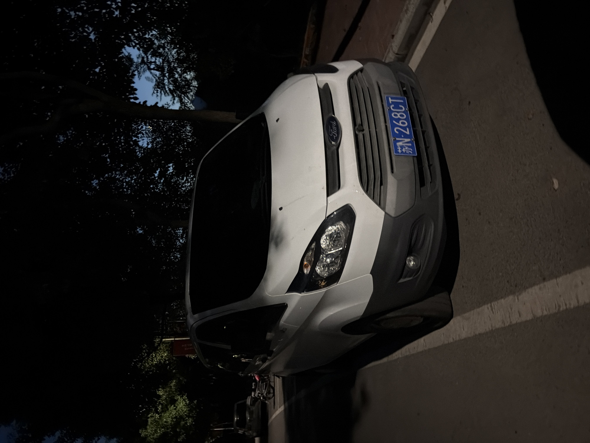 In the city of Jiangjin, China, at dusk, a white Ford Transit van is parked on the side of a road. The van is facing forward, and its headlights are on. The van has a gray bumper and a license plate that reads "苏N-268CT". The road is paved with asphalt and has a white line marking the lane. Trees with dark green leaves are visible on the left side of the road. In the background, a dark sky with some visible clouds suggests the setting sun. The van appears to be unoccupied.