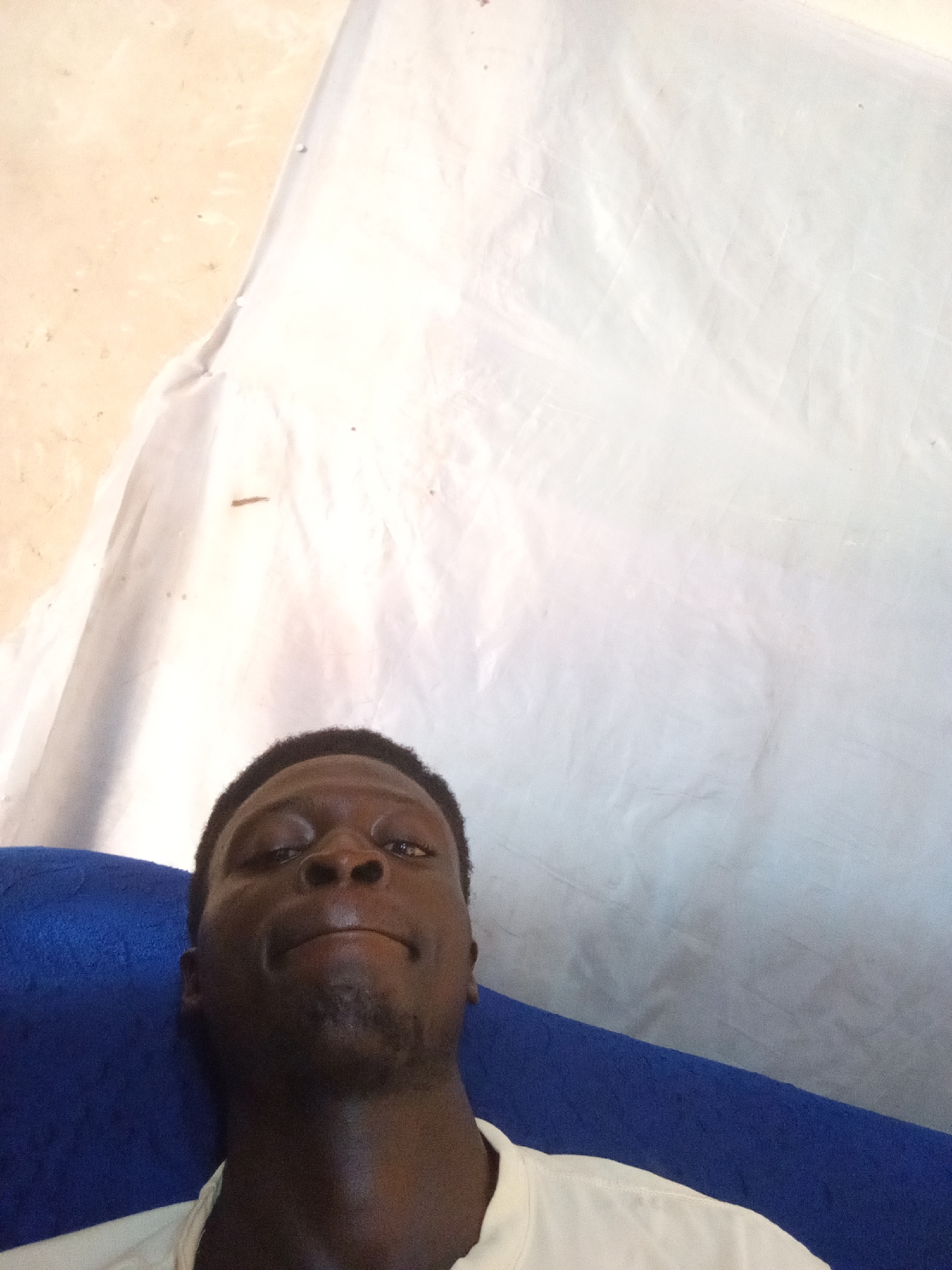 In this selfie, a young Black man with dark skin and short, dark, curly hair is lying down on a blue surface. He is looking directly at the camera with his mouth slightly open and his eyes looking upward. He is wearing a white t-shirt. Behind him, a large, white sheet is draped, creating a textured backdrop. The sheet appears to be stretched and creased, with some areas appearing lighter and others darker due to the lighting and folds. To the left of the sheet, a textured surface with a light brown or beige hue is visible, with small dark specks. The overall scene is dimly lit. The image does not contain any information that indicates the location within the city of Nyabe, Nigeria.