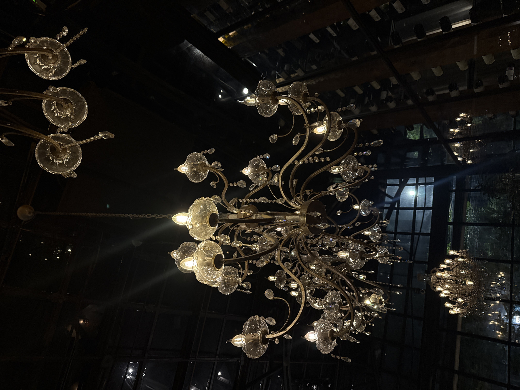 The image shows a low-angle view of ornate chandeliers, presumably indoors. The chandeliers are a combination of clear glass and metal frames. The background is dark, with windows visible in the distance. The overhead ceiling reveals wooden support beams and a mirrored surface. Bottles are stowed above the wooden beams. The location is Mexico City, Mexico.