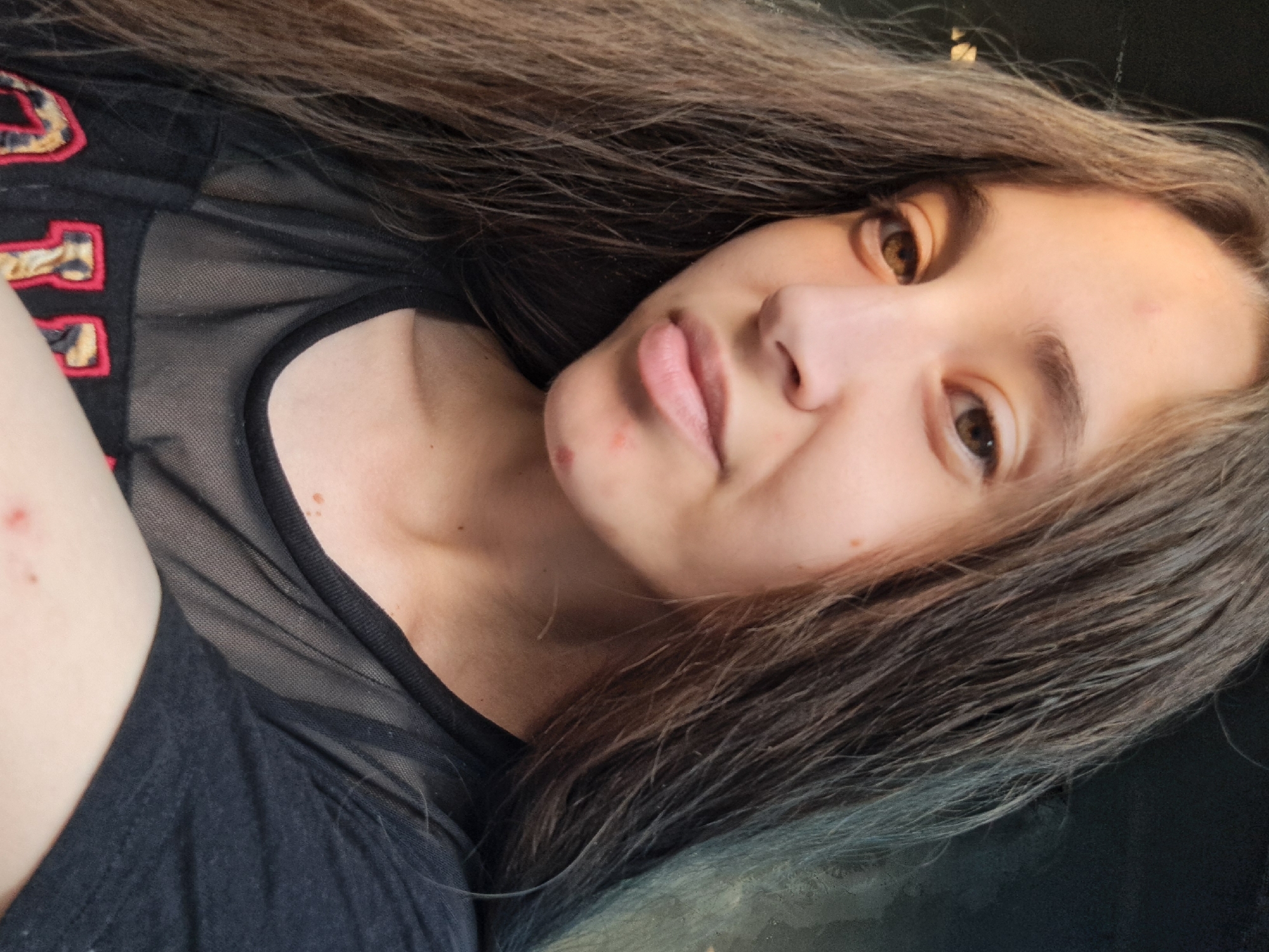 The image is a close-up, waist-up, selfie-style photograph of a young woman with long, straight, dark brown hair. She is facing the camera, with her head slightly tilted. Her eyes are brown, and she has a neutral expression. There are a few small blemishes visible on her face and chin. She is wearing a black t-shirt with sheer netting on the upper chest area and red and leopard print lettering on the lower part of the shirt. The background is dark and indistinct, suggesting an indoor setting or a dark outdoor environment. There are no discernible environmental details that would suggest a specific location within the city of Vypasne, Ukraine.