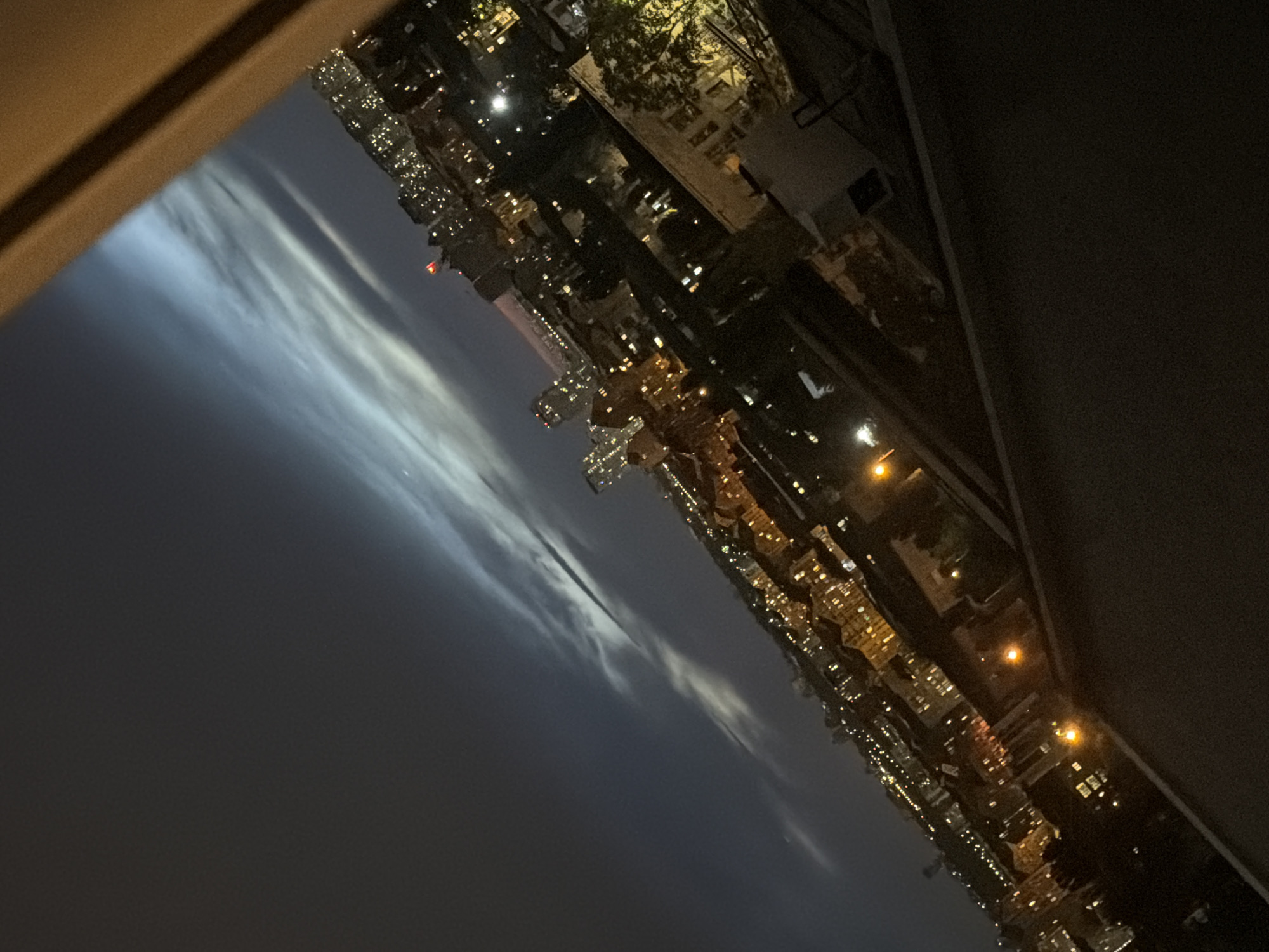 An aerial view captures a night scene over a city, likely Kyiv, Ukraine, with buildings illuminated by artificial lights. The perspective is angled, suggesting the photograph was taken from an elevated position, possibly a balcony or window. Wispy clouds streak across the dark night sky, reflecting ambient light, creating a soft, ethereal glow. The cityscape is dense with structures of varying heights, some featuring prominent lighted windows, indicating residential or commercial activity. Streets and pathways are visible, lined with streetlights and the glow from buildings. The overall impression is a tranquil, urban panorama under a vast, cloudy sky.