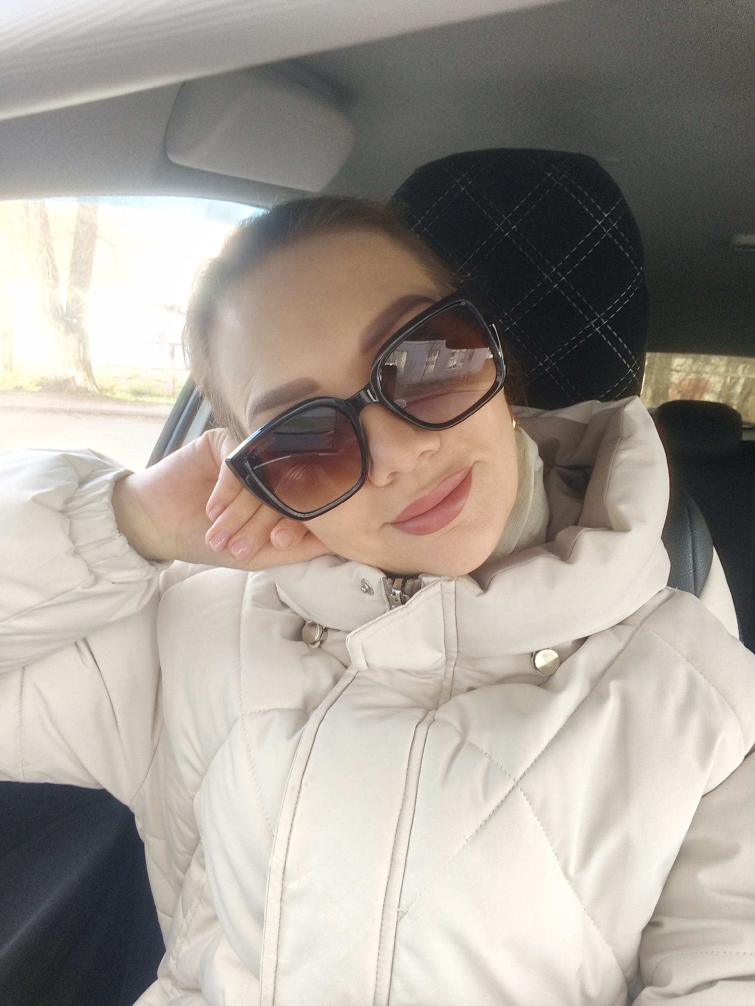 The image is a bright, eye-level selfie of a woman seated in a car. She is looking directly at the camera with a soft smile, her head tilted slightly, and her right hand resting gently on her cheek. She wears oversized, dark-framed sunglasses, which reflect a bright outdoor scene and a building. The text "P.PREMIUM" is faintly visible on the arm of her sunglasses.
The woman is dressed in a light, likely cream or off-white, quilted puffer jacket with a high, protective collar, under which a light-colored turtleneck or scarf is visible. Her hair is pulled back, and her eyebrows are well-defined. Her manicured nails are painted with a subtle, light pink polish.
The scene is set inside a car, indicated by the light-colored headliner and a sun visor above her. Behind her, a black car seat headrest with a distinctive quilted pattern and white stitching is visible. Through the car window to her left, bright sunlight illuminates bare trees, suggesting a clear, sunny day in a cooler season like late autumn or winter. The overall impression is one of casual comfort and relaxation.