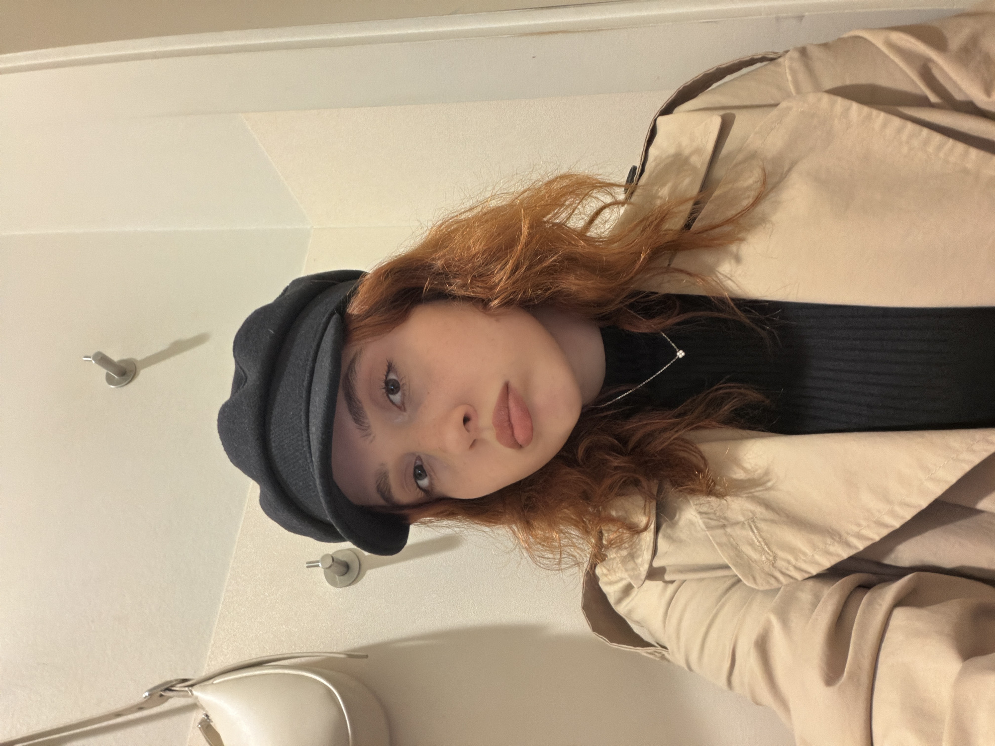 A young, fair-skinned woman with red curly hair, wearing a black sweater, a beige trench coat, and a black hat, is looking at the camera. She has a necklace on. The background consists of a white wall with two metal hooks. Part of a white purse is visible in the lower left corner. The photo appears to be taken indoors. The location is impossible to determine from the photograph.
