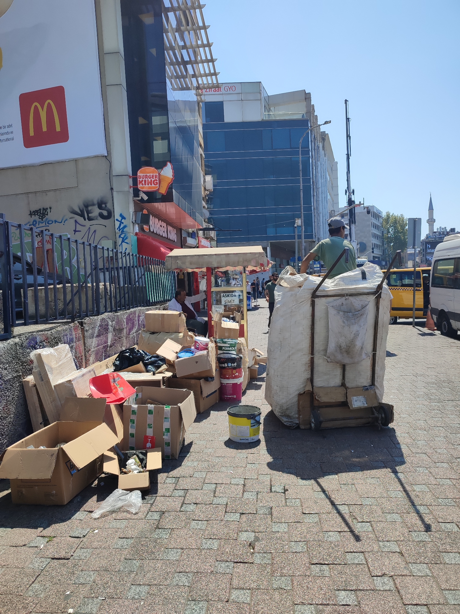 In Kadıköy, Turkey, on a sunny day, a man wearing a grey cap and a green t-shirt stands on a cart laden with a large, white, sack-like bundle. The cart has a metal frame and two wheels. The man is in the process of moving or has just finished moving the cart. In the foreground, numerous cardboard boxes are piled on the cobblestone pavement, some open and others sealed. Several buckets, including one yellow and one white, are also present.
Behind the cart and boxes, a street vendor's stall with a red awning is visible. Further back, the architecture of the city is evident, with modern buildings featuring glass facades and older structures. A prominent McDonald's advertisement is plastered on a large white surface on the left, with a red square containing the iconic golden arches logo. Adjacent to it is a Burger King sign. The scene suggests a bustling urban environment with commercial activity and street-level operations. A yellow taxi and a white van are also parked on the street in the background. The sky is clear and blue, indicating fair weather.